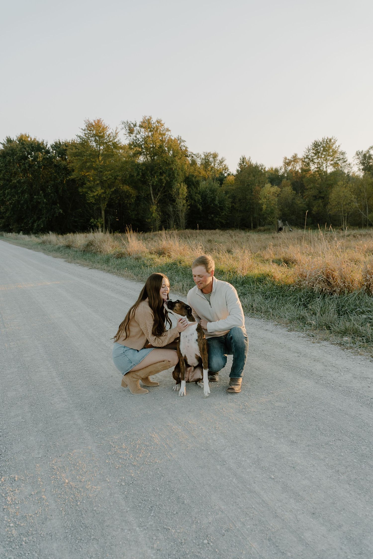 Young couple crouching on a rural gravel road petting their dog at sunset with trees and fields in background.