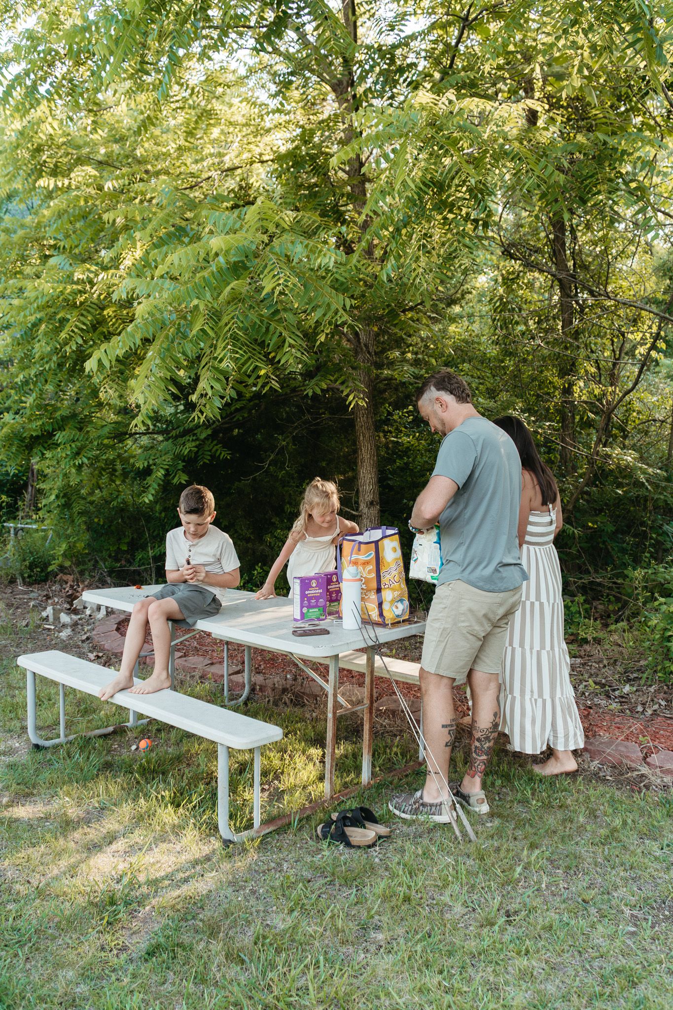 A family unpacks snacks at an outdoor picnic table surrounded by lush green trees in summer.