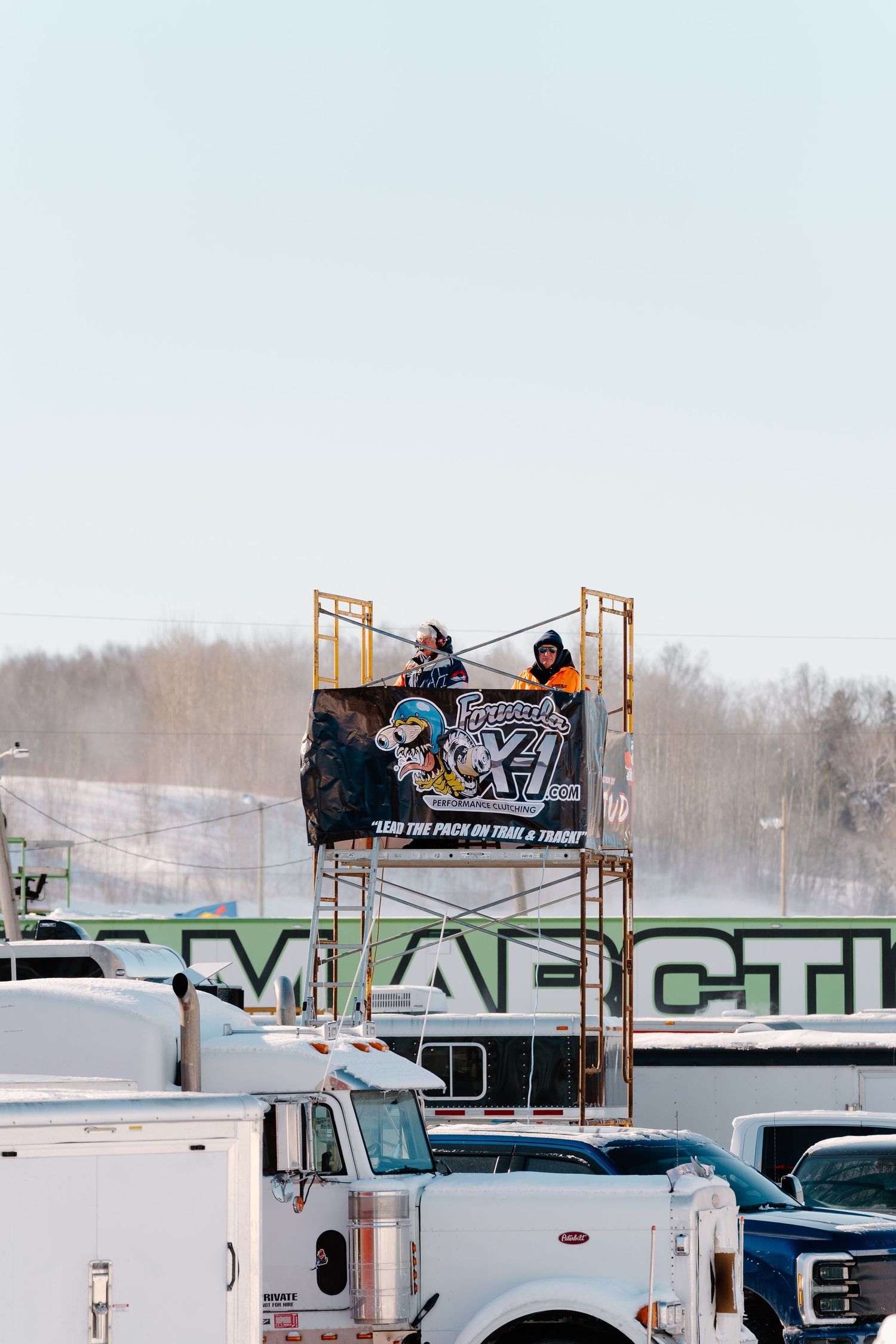 Two people standing on a scaffolding tower with a Polaris racing banner at a winter snowmobile event.