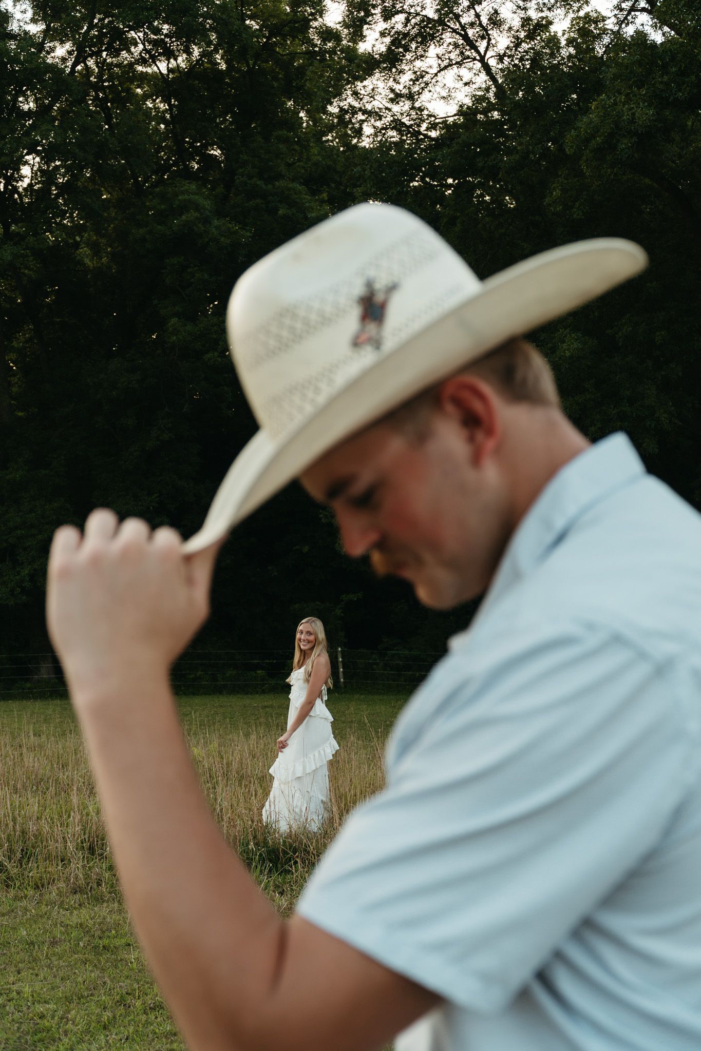 Man in cowboy hat tipping brim with woman in white dress in background.