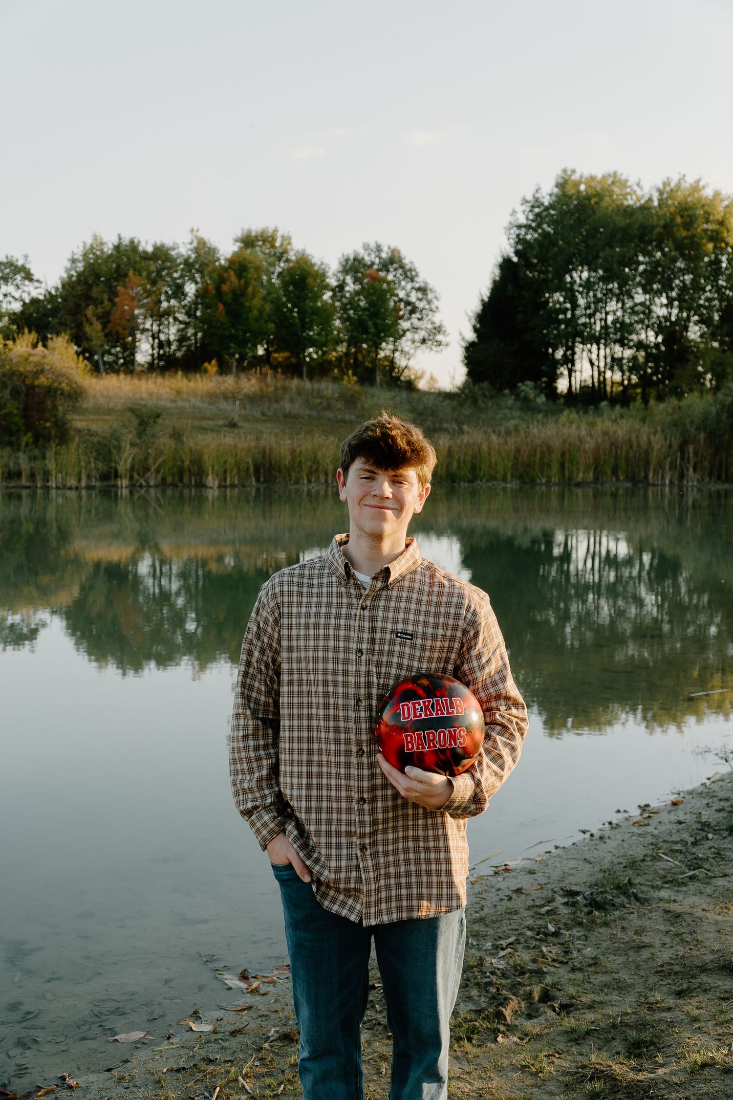 Young man holding a basketball standing by a calm reflective pond surrounded by autumn trees.