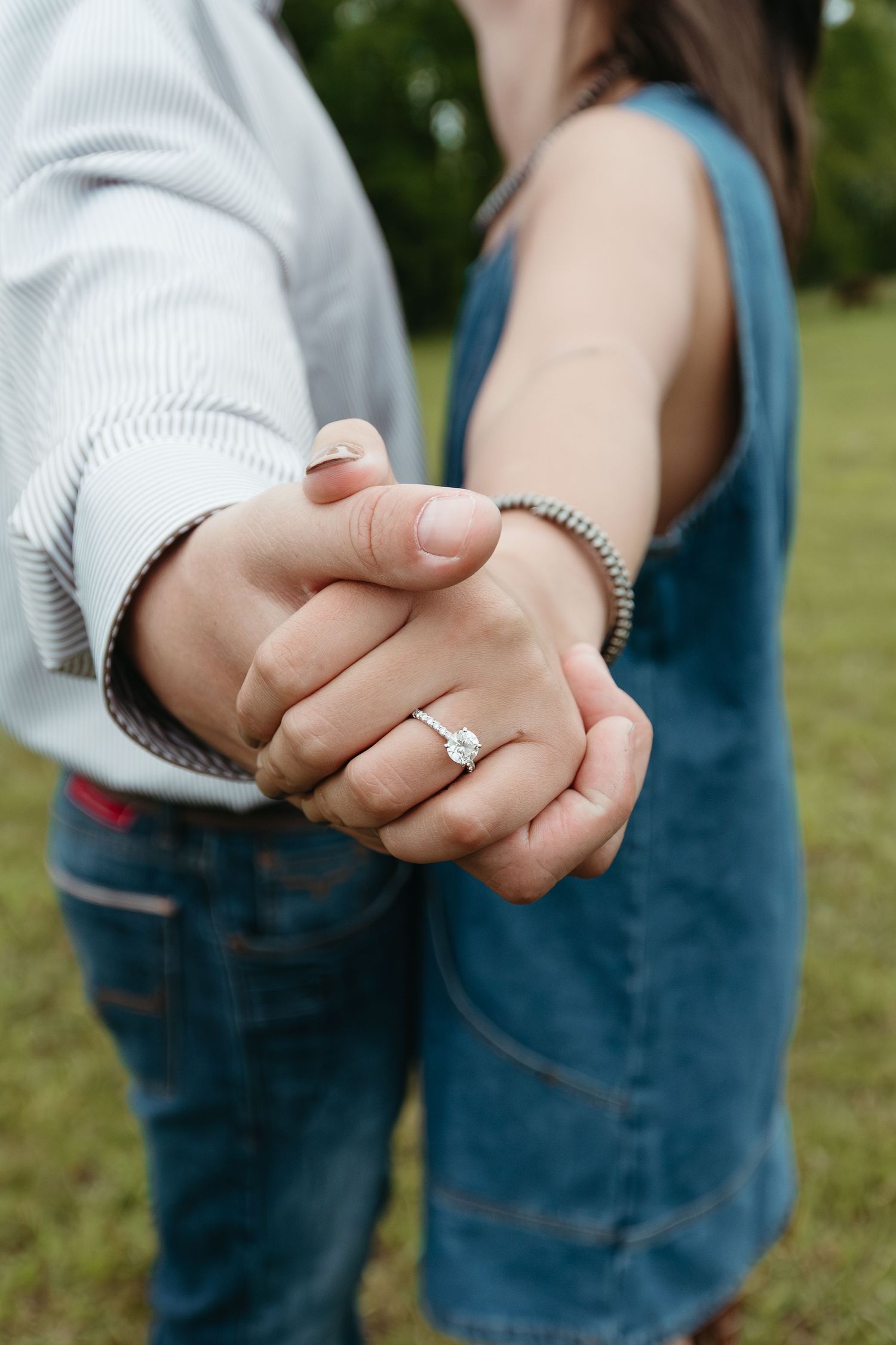 Close-up of couple holding hands showing diamond engagement ring and beaded bracelet outdoors.