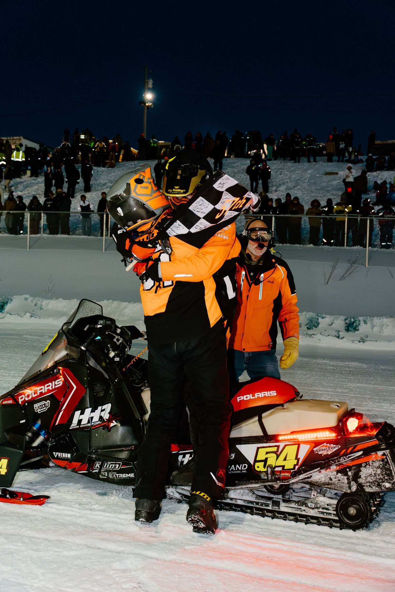 Snowmobile racer number 54 on a Polaris sled celebrates victory with a checkered flag at a night race.