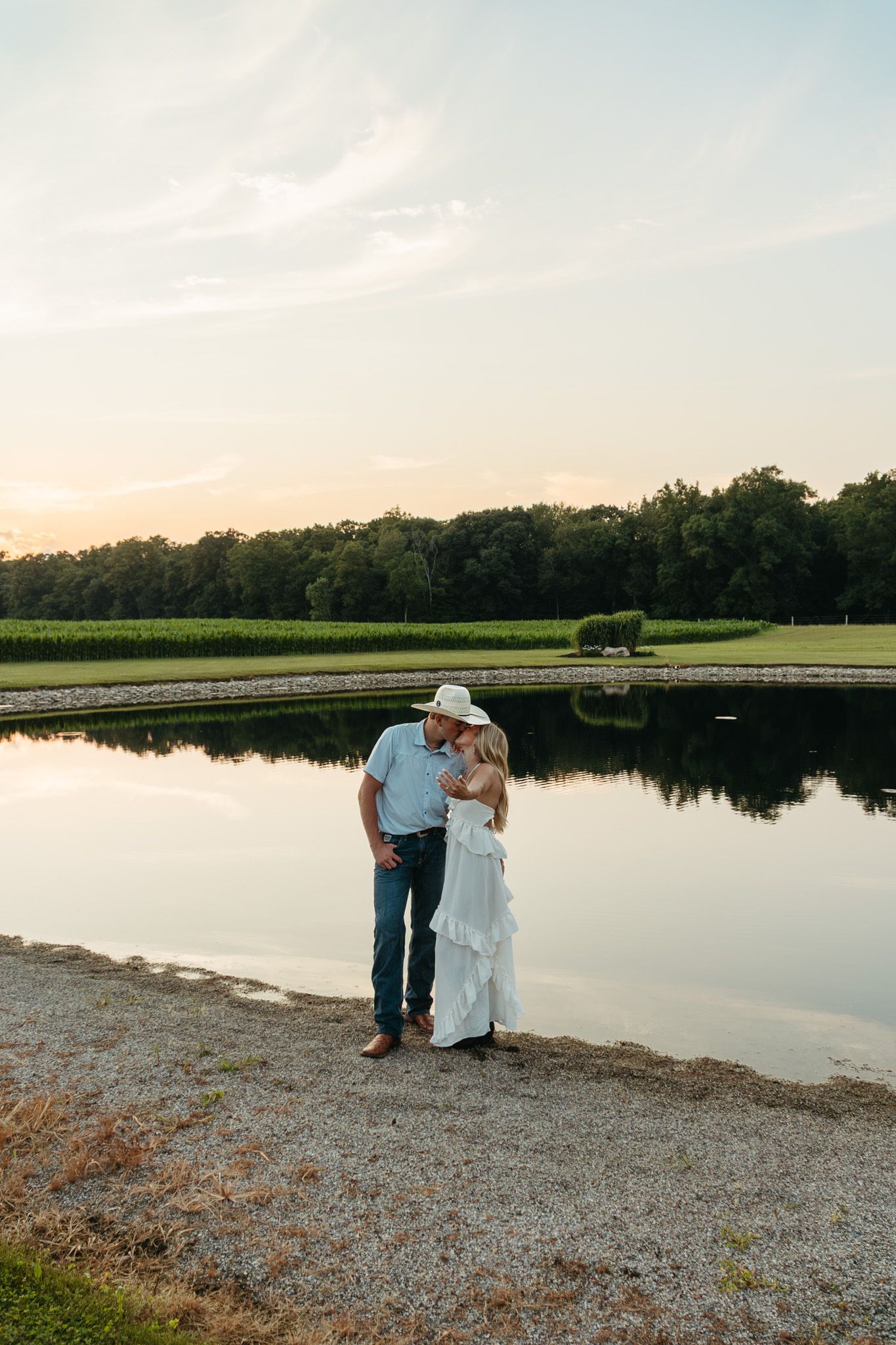 Couple sharing a romantic kiss beside a calm reflective pond at a beautiful farm sunset.
