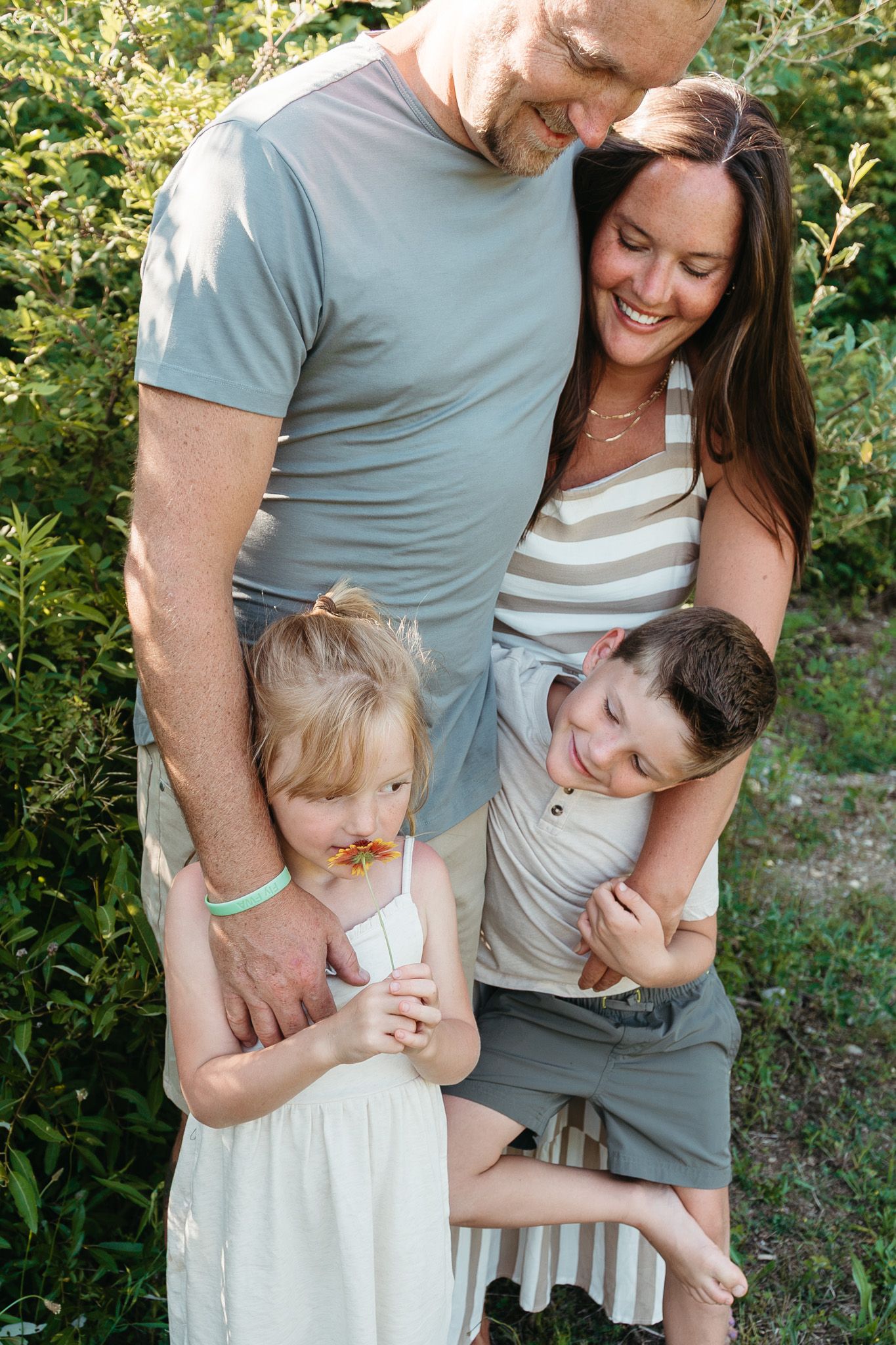 Happy family of four smiles together outdoors surrounded by green bushes in warm sunlight.