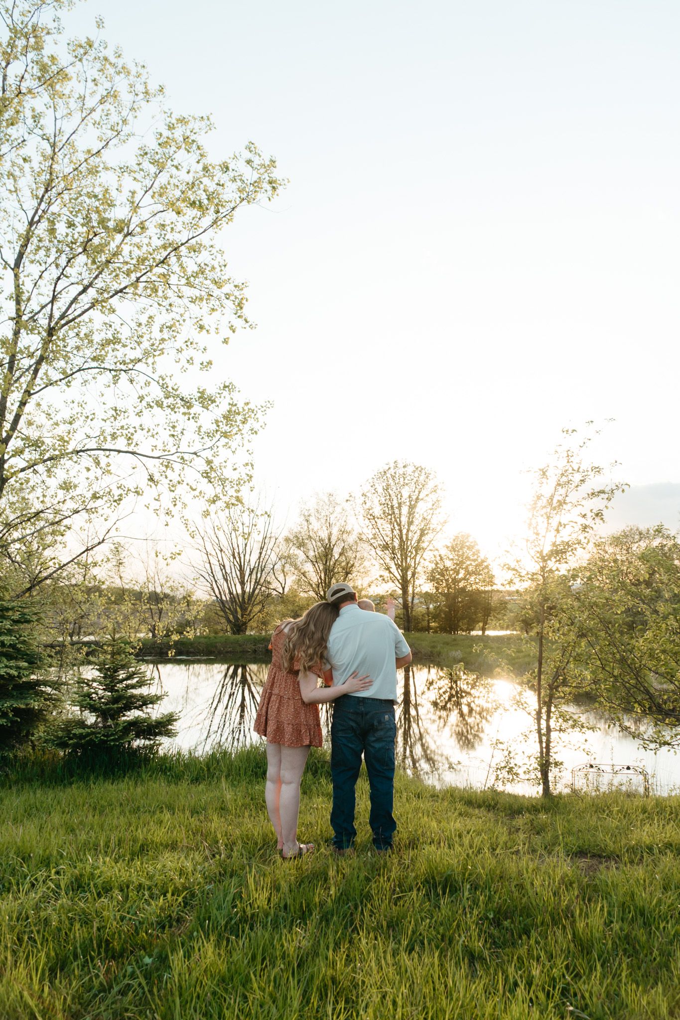 Couple standing arm in arm at golden hour overlooking a serene pond surrounded by lush green trees and grass.