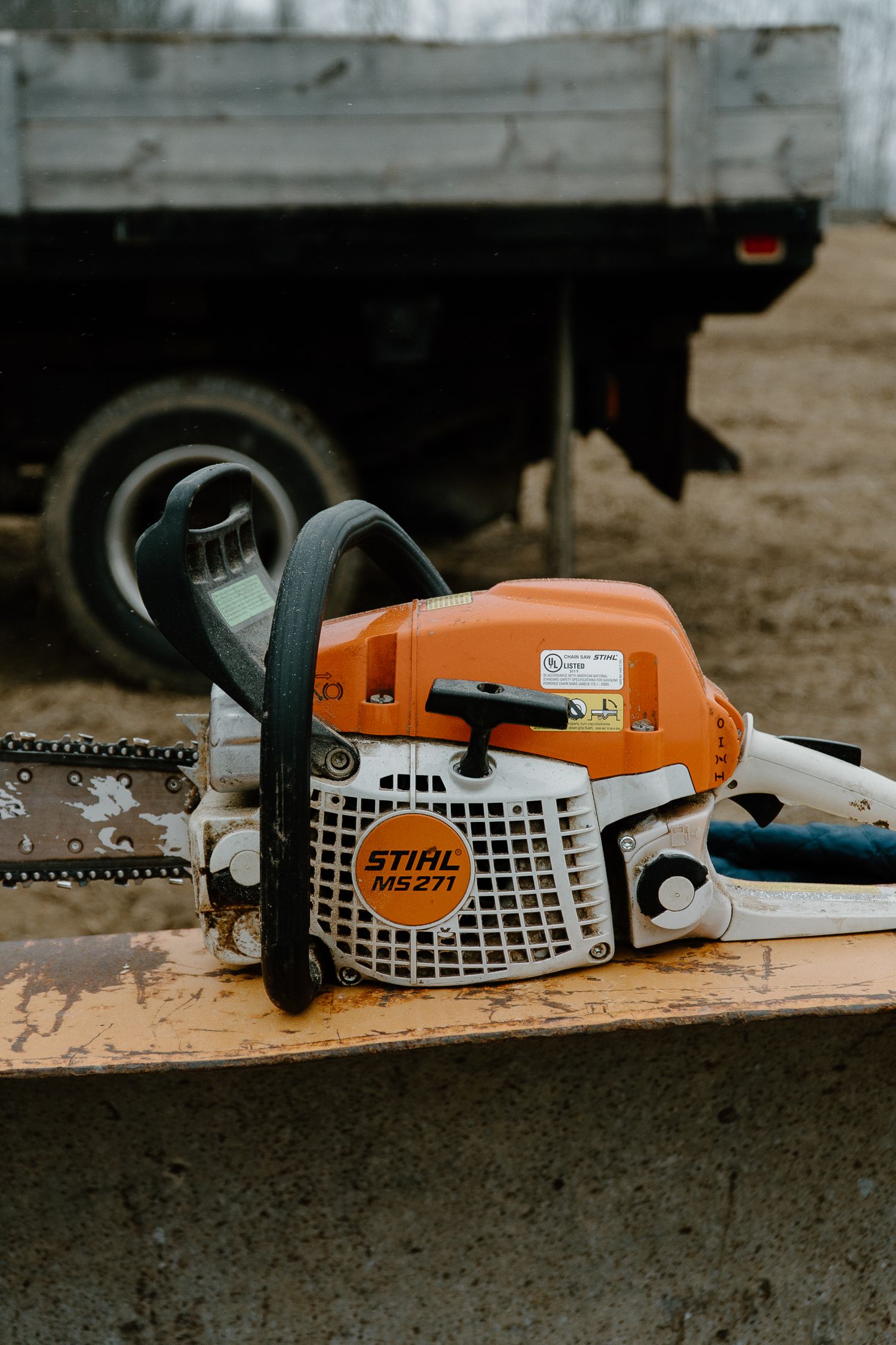 Stihl MS271 chainsaw resting on a wooden surface with a truck in the background on a dirt lot.
