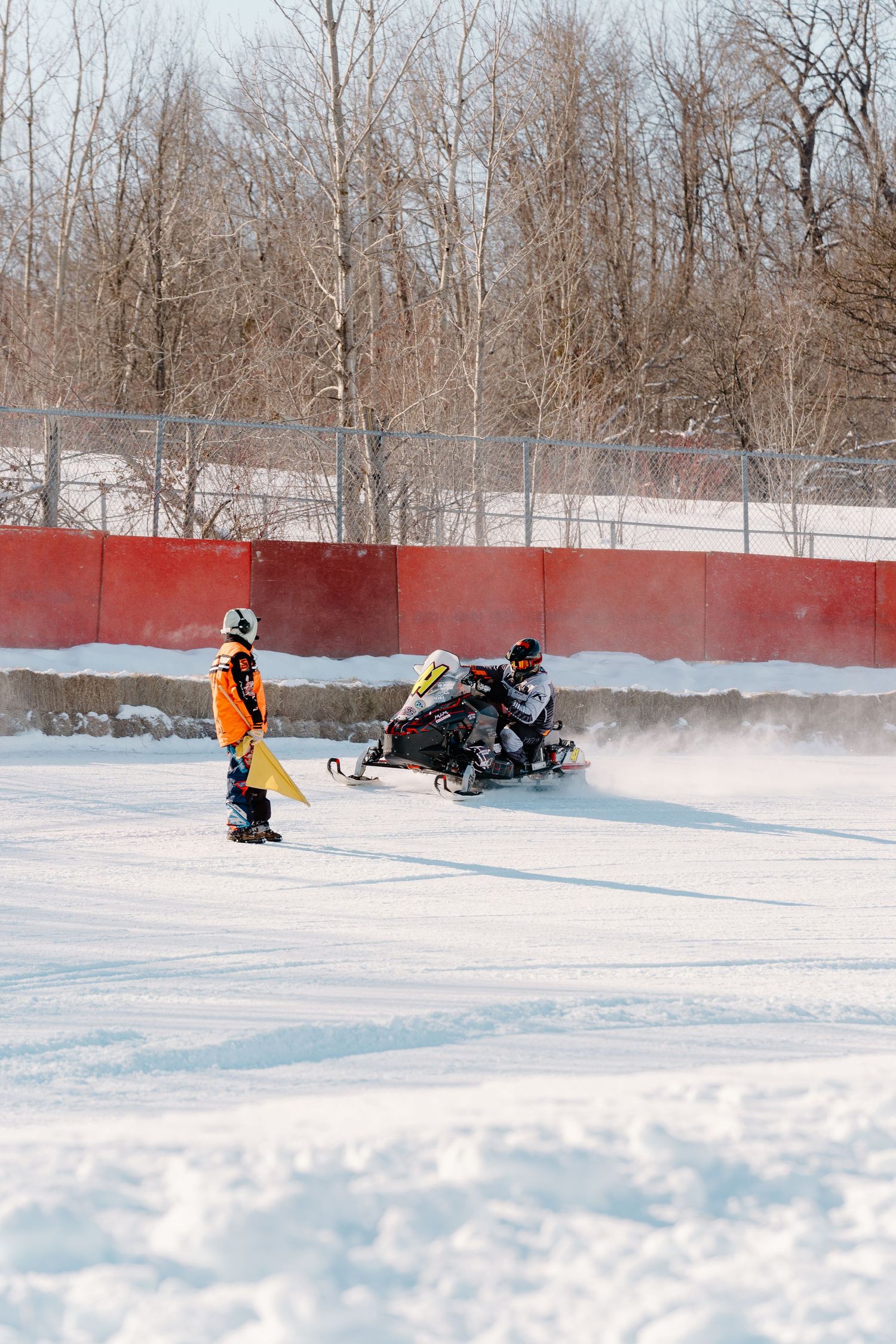 Race official in orange jacket holding yellow flag near a snowmobile racer on a snowy winter track.
