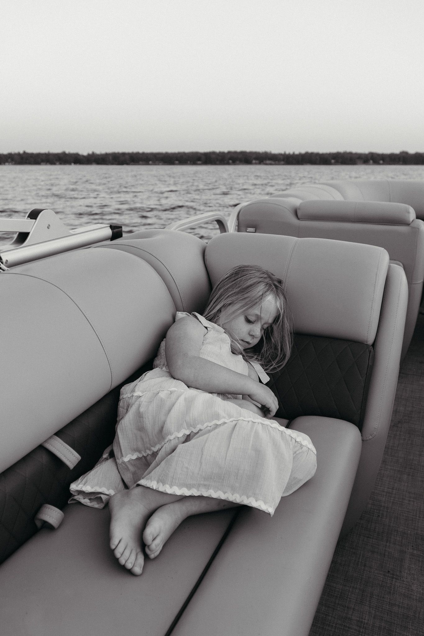 Black and white photo of young girl in white dress sleeping curled up on pontoon boat seats.