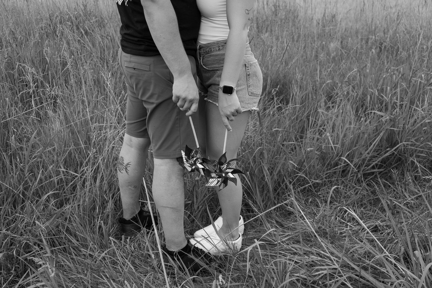Black and white photo of couple holding wildflowers while standing together in tall grass field.