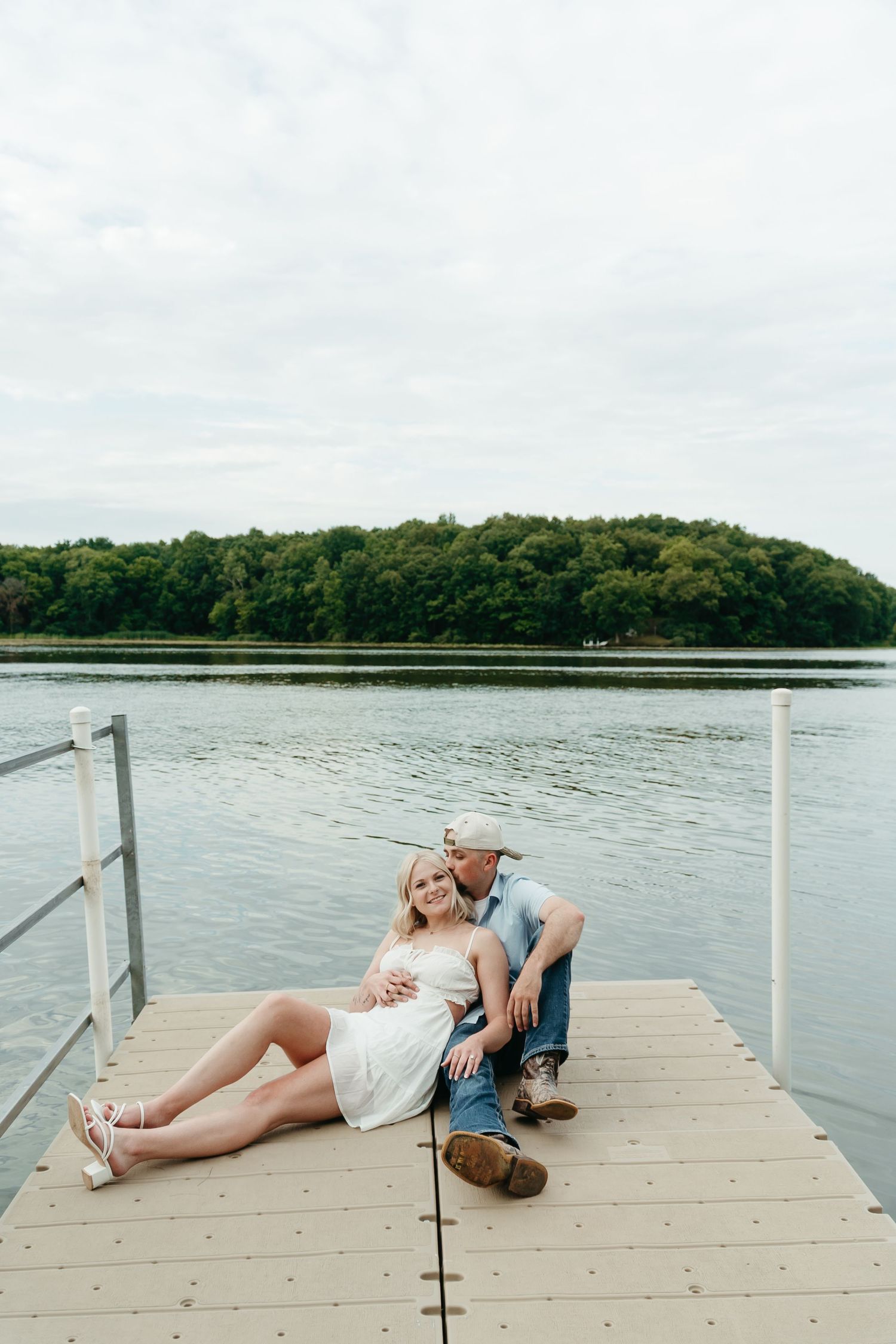 Couple relaxing on a lakeside dock, man kissing woman in white dress with tree-covered island in background.