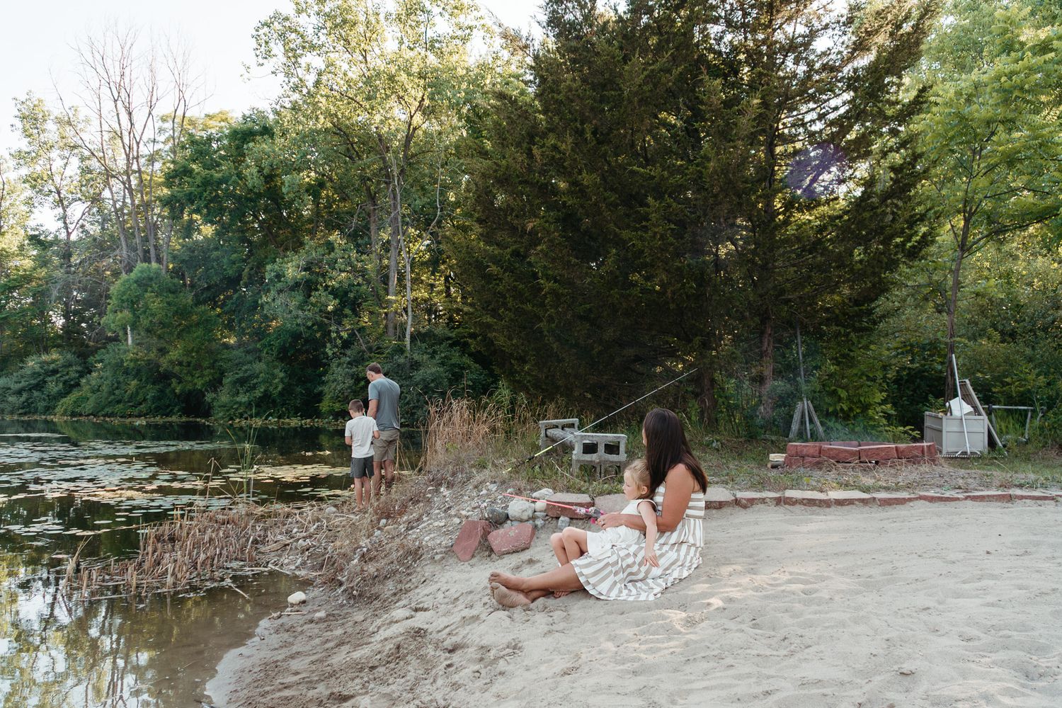 A woman sits on a sandy riverbank while another person stands near the water surrounded by trees.
