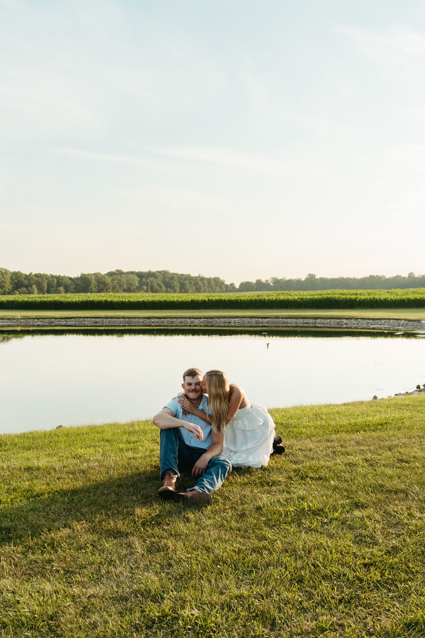 Couple sitting close together by a reflective pond on green grass during a warm sunny day.