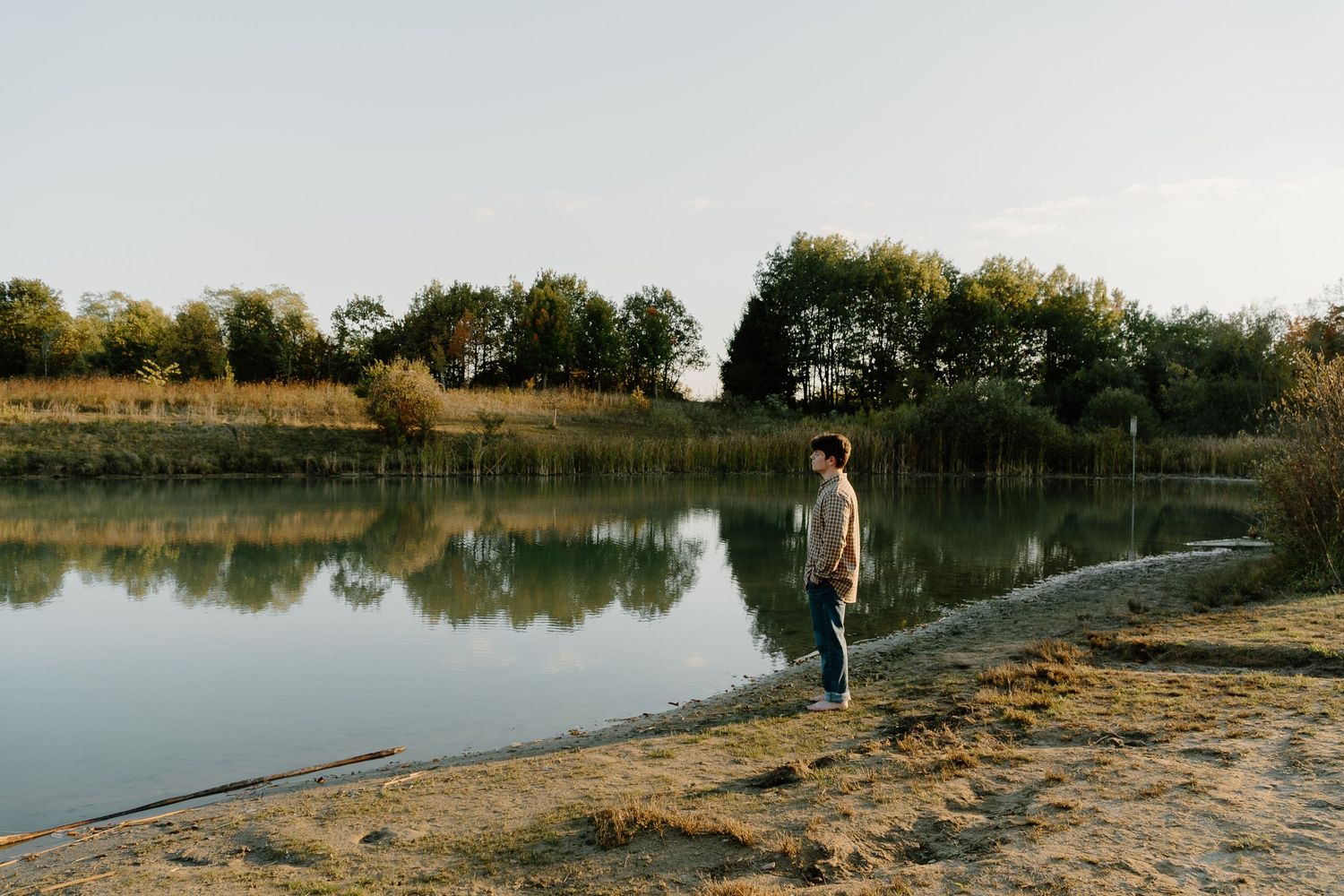 Young man standing alone by a peaceful reflective lake surrounded by trees on a clear day.