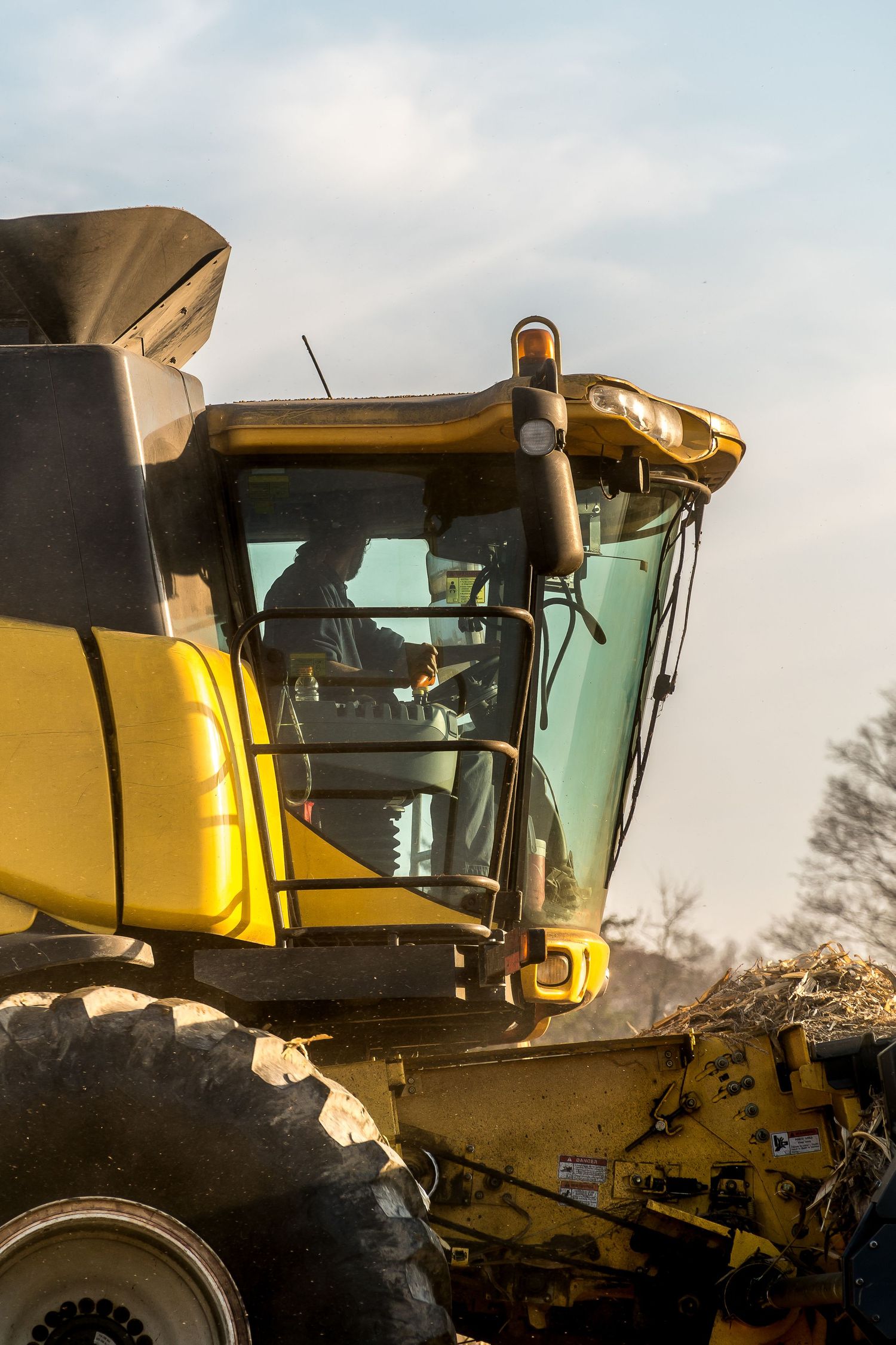 Farmer operating a yellow New Holland combine harvester cab during autumn crop harvest at golden hour.