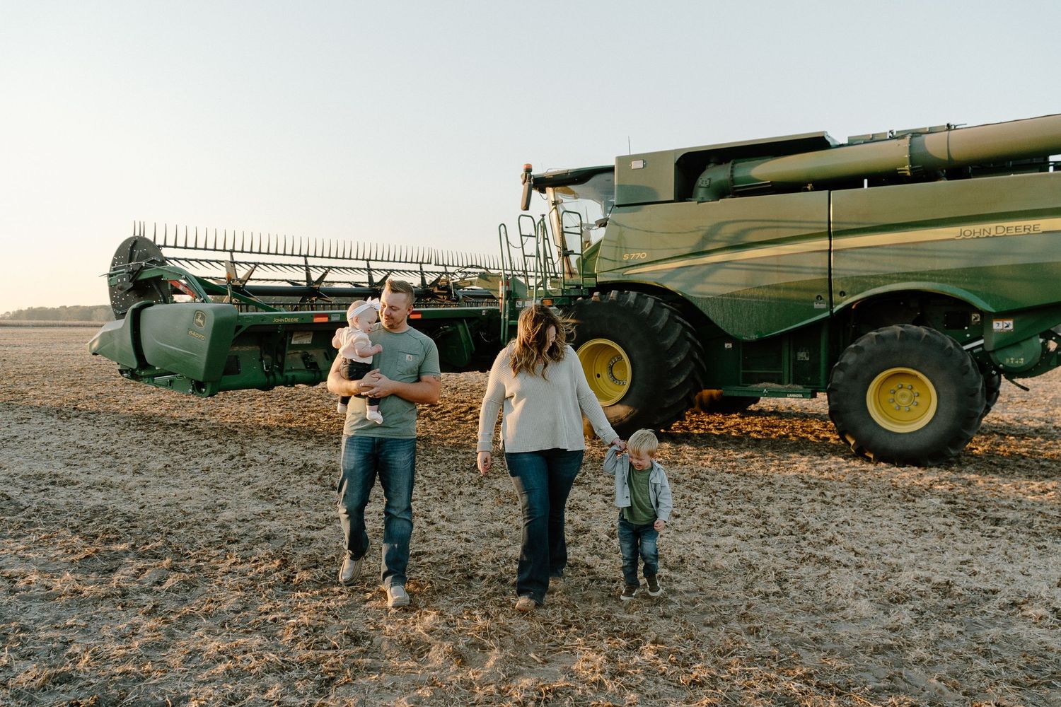 Young farm family walking together in front of a green John Deere combine harvester at sunset.