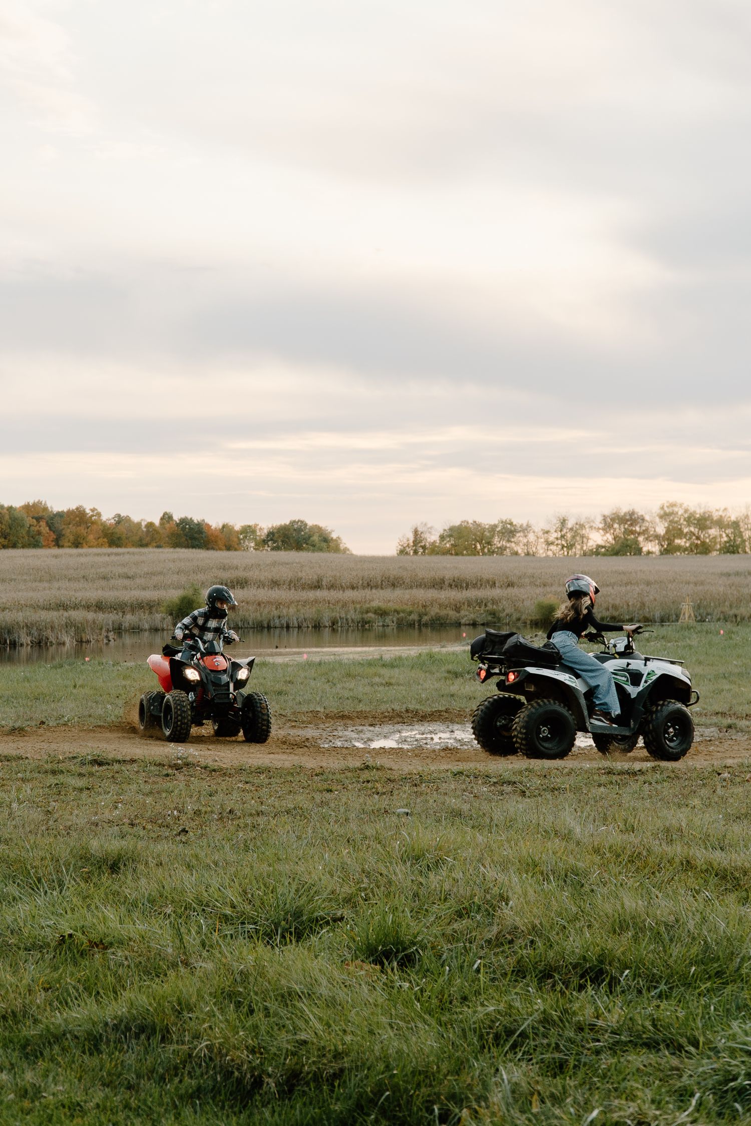 Two riders on ATVs racing across a muddy field on a cloudy day.