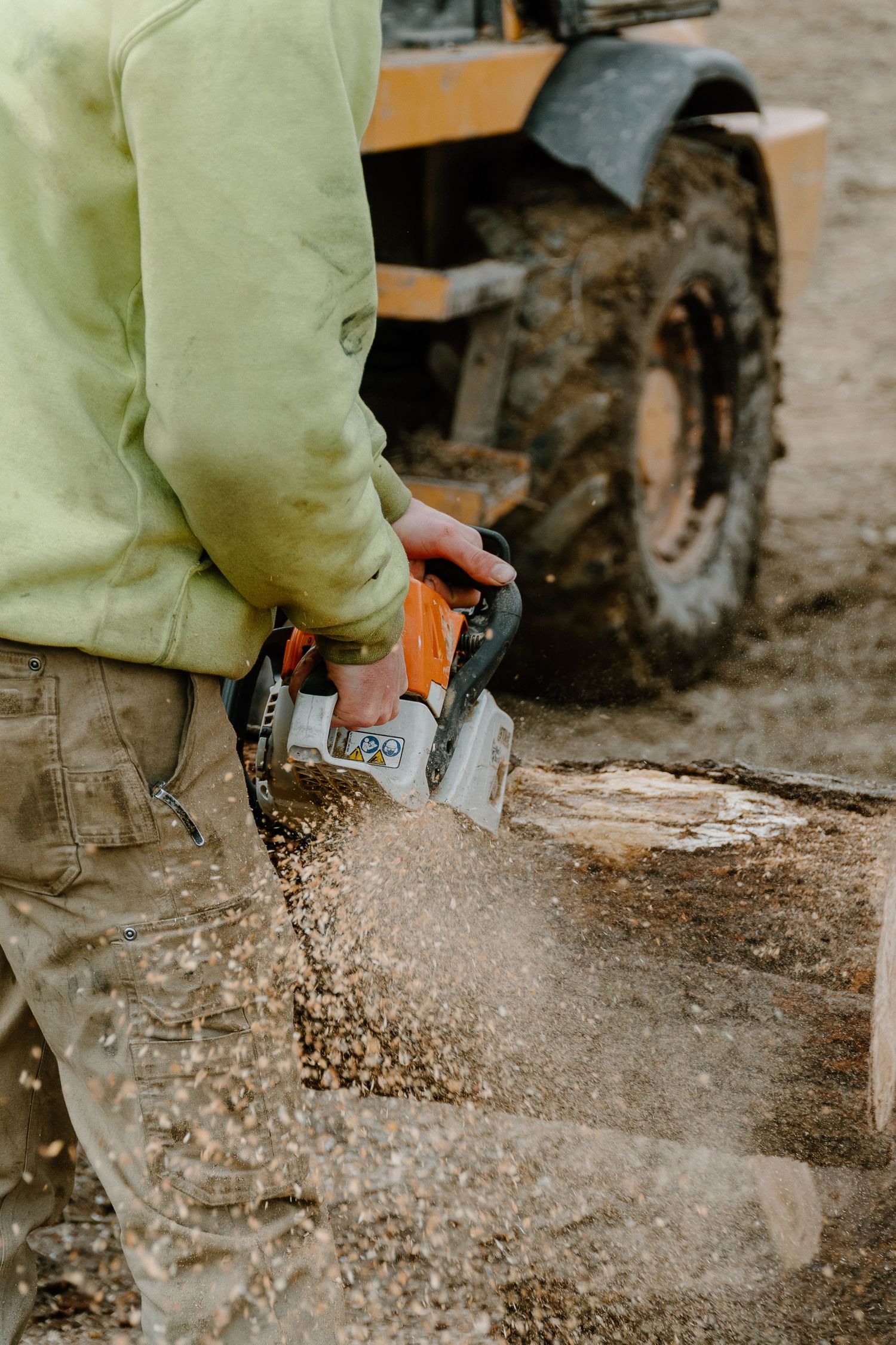 Worker using an orange chainsaw to cut a log with sawdust flying, heavy machinery in background.