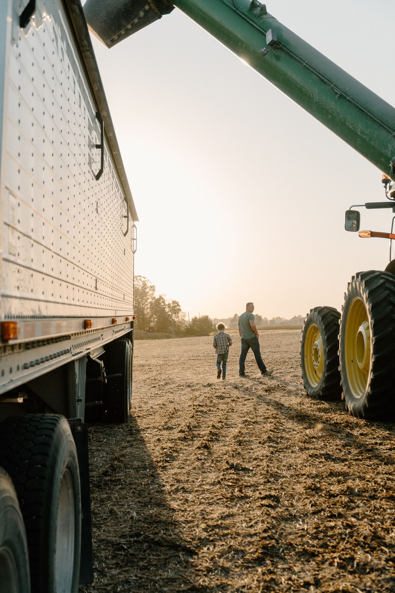 Farmer and child standing between a grain truck and tractor in a harvested field at sunset.