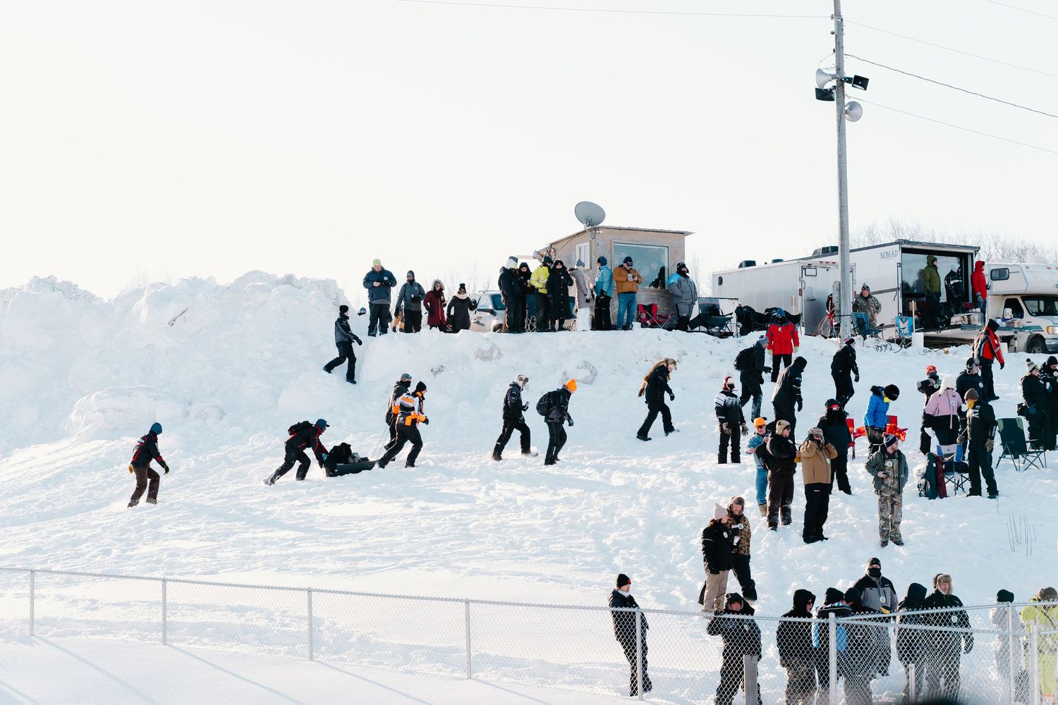 A crowd of people play and walk on a large snow hill during a sunny winter day.