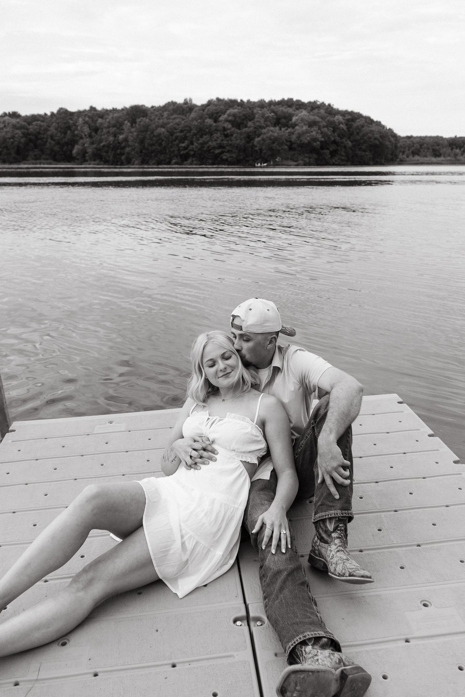 Black and white photo of couple relaxing on a wooden dock by a serene lake with forested island.