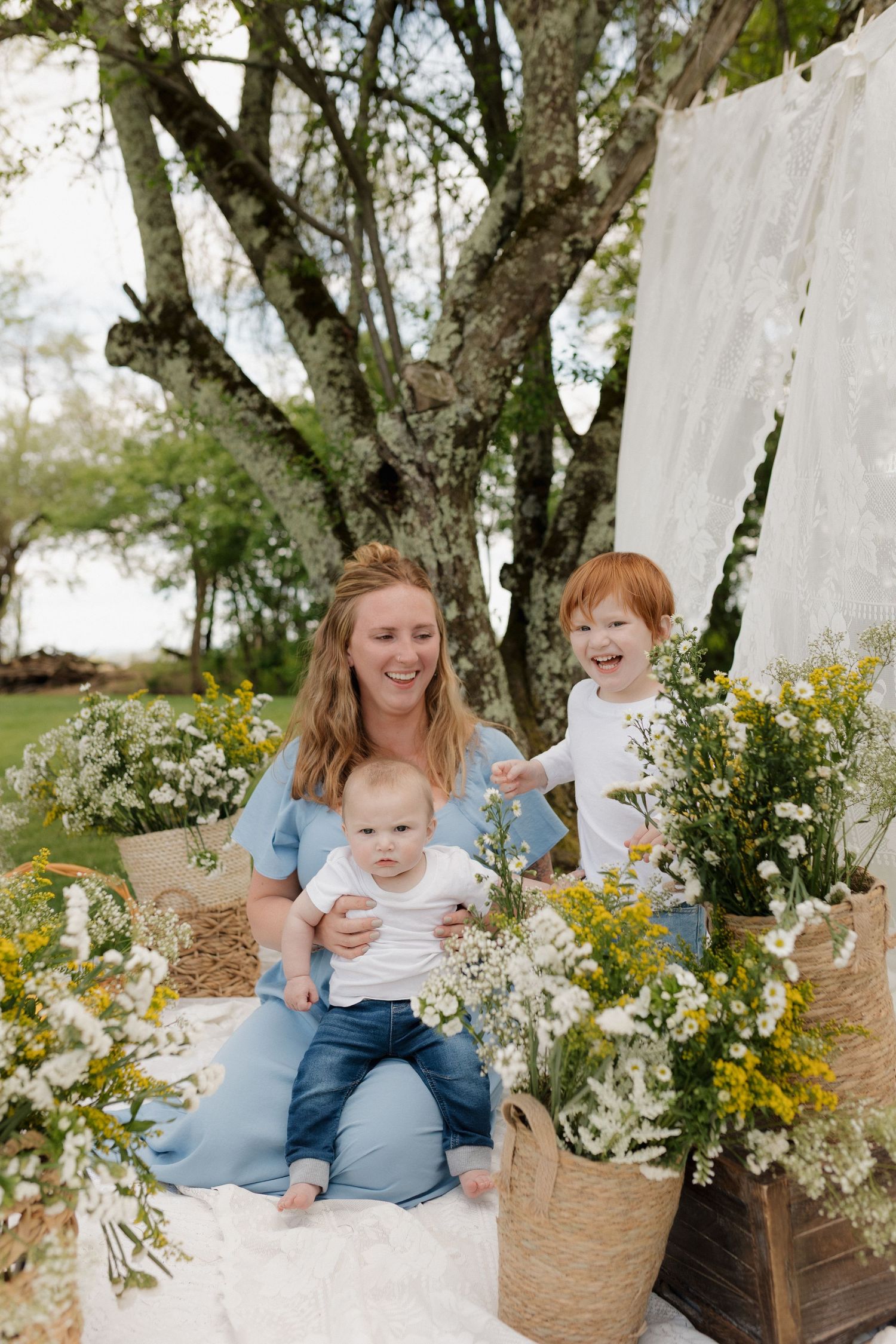 A smiling mother in a blue dress sits outdoors with her baby and toddler surrounded by yellow and white wildflowers.
