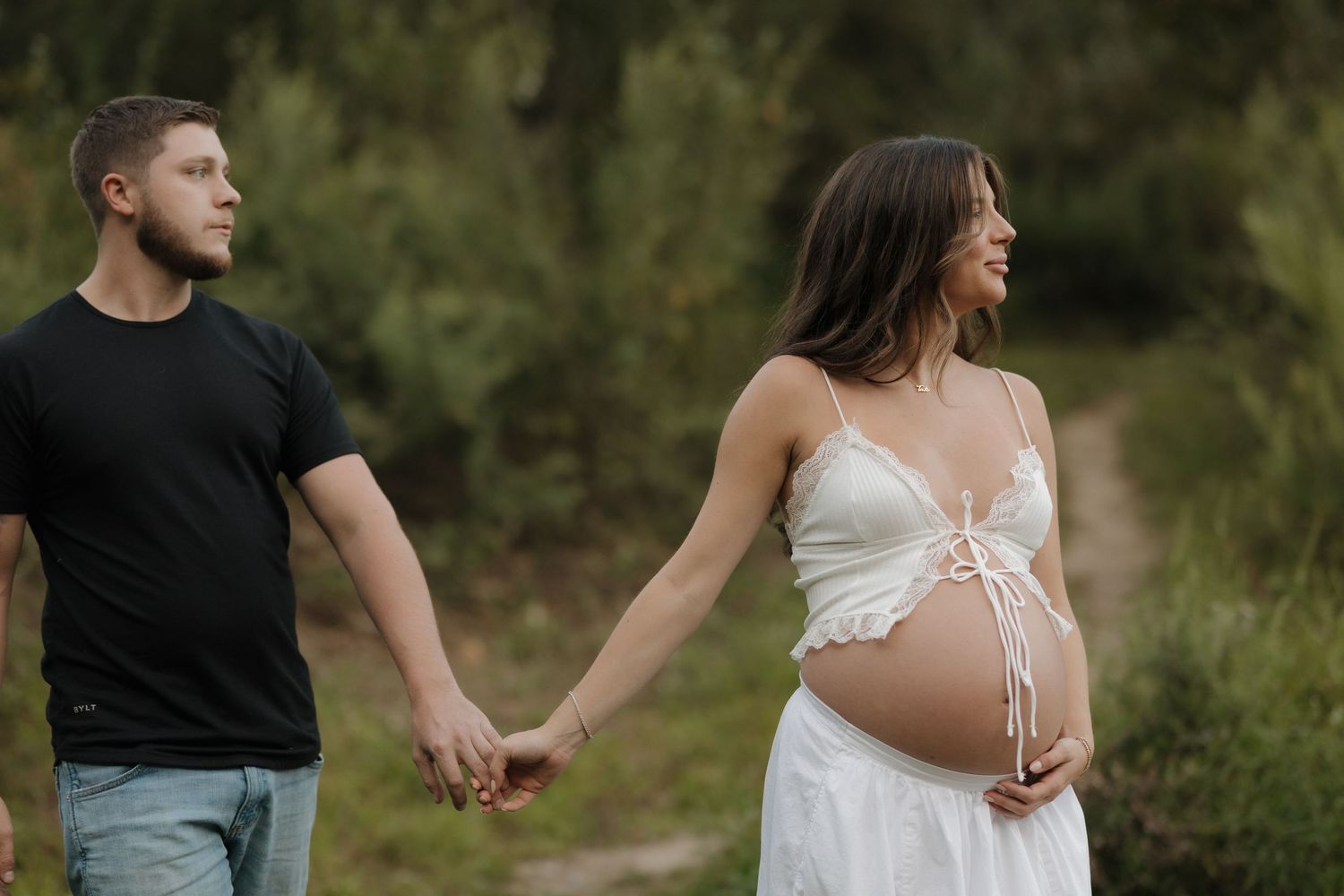 A couple holds hands while walking outdoors, with a person wearing a flowing white maternity dress.