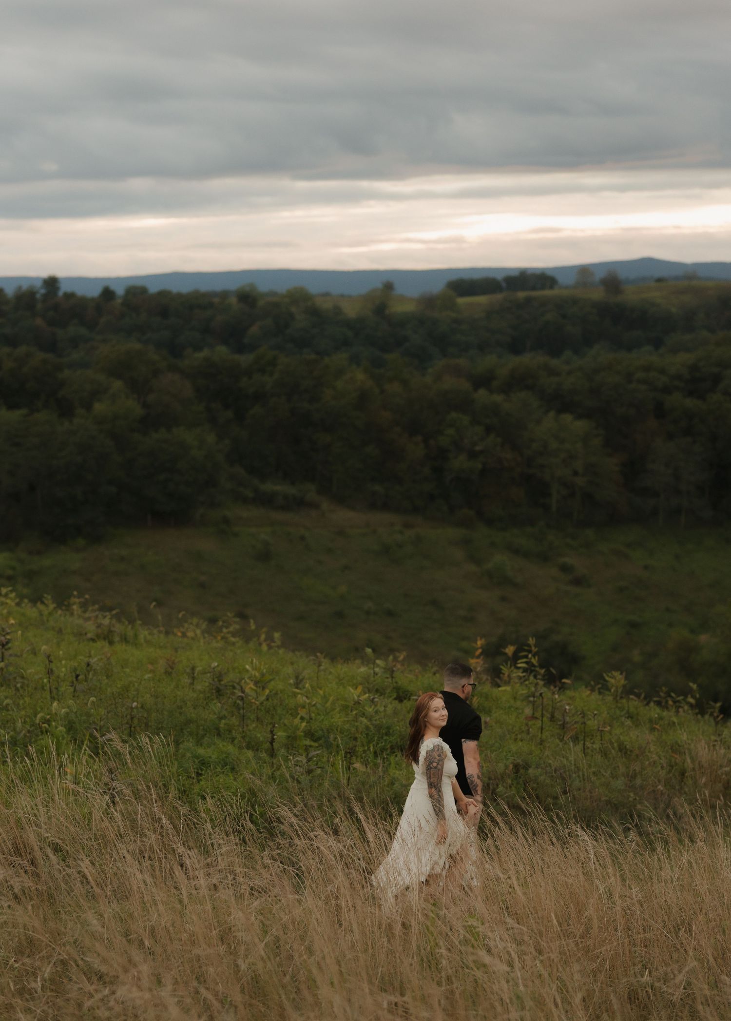 Couple standing in tall grass field against dramatic mountain sunset backdrop.