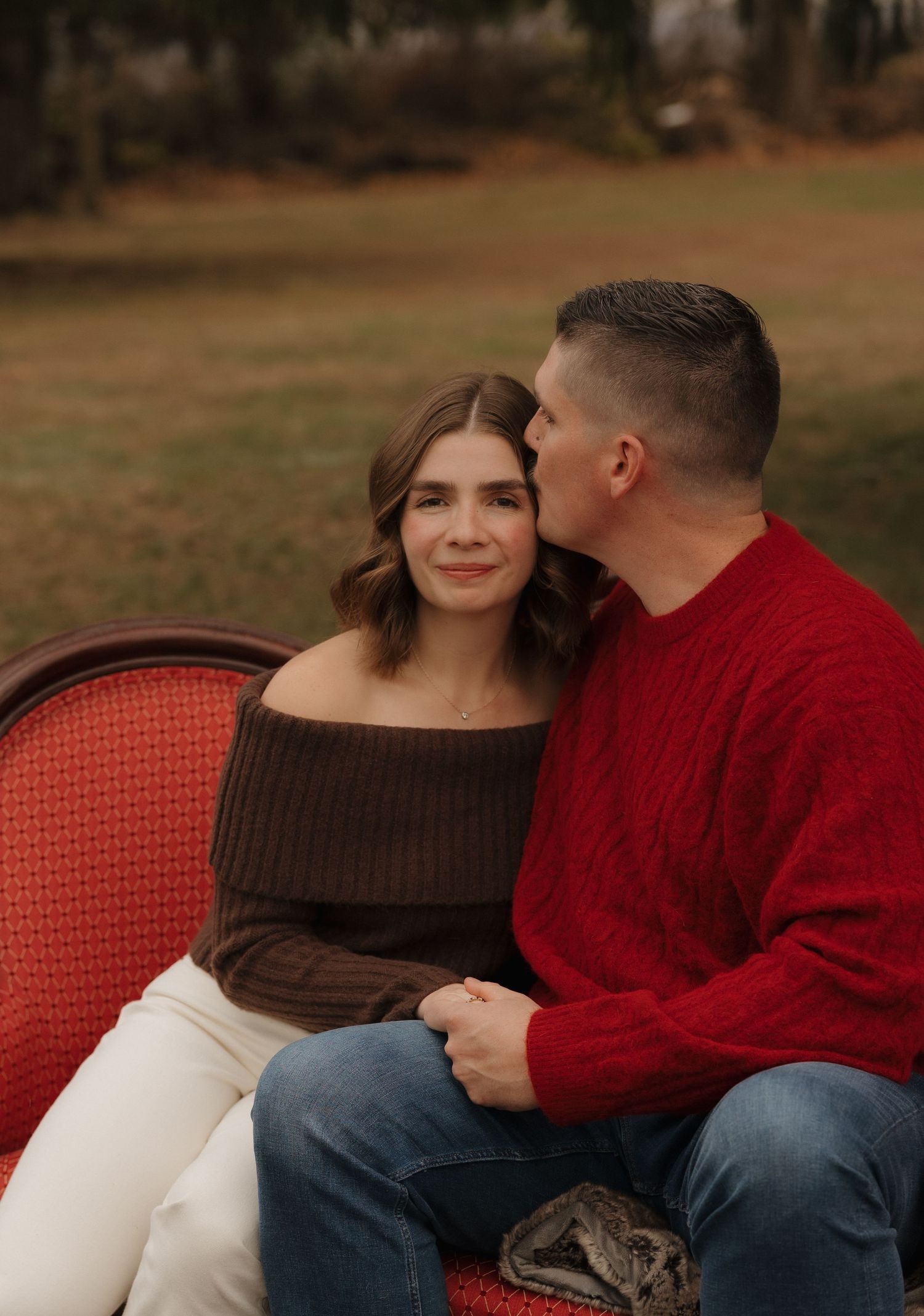 Couple sits together on a red couch outdoors during an autumn photoshoot.