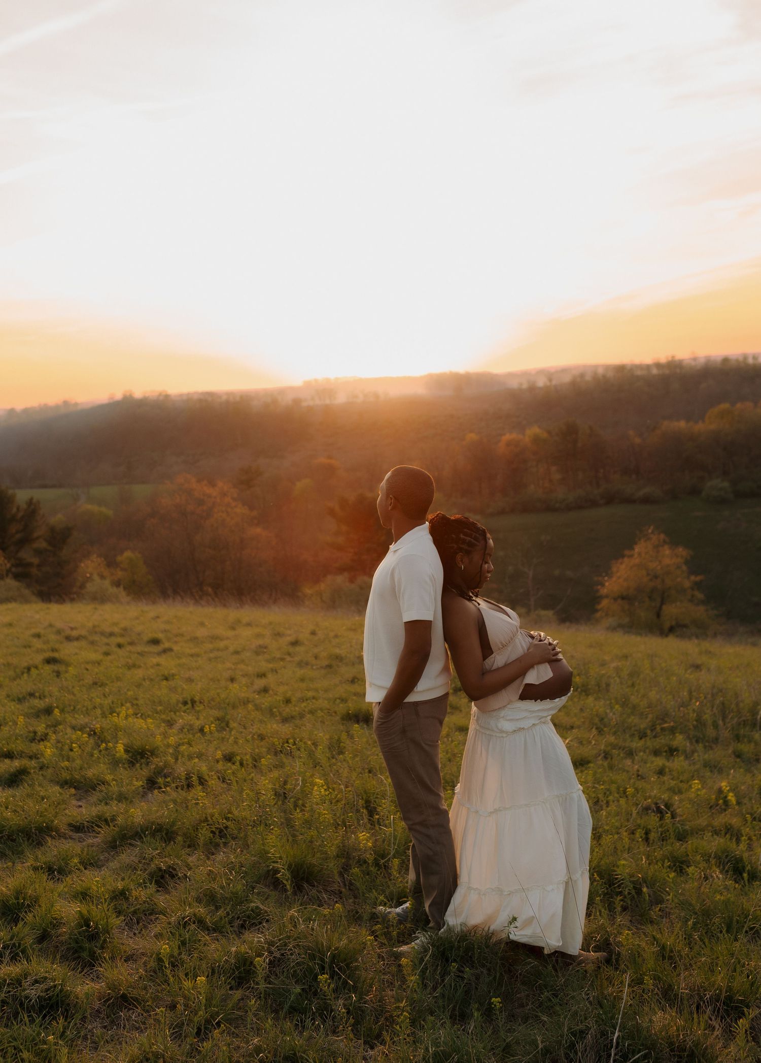 A couple embraces in a golden field at sunset, the woman in a white dress, surrounded by rolling hills.