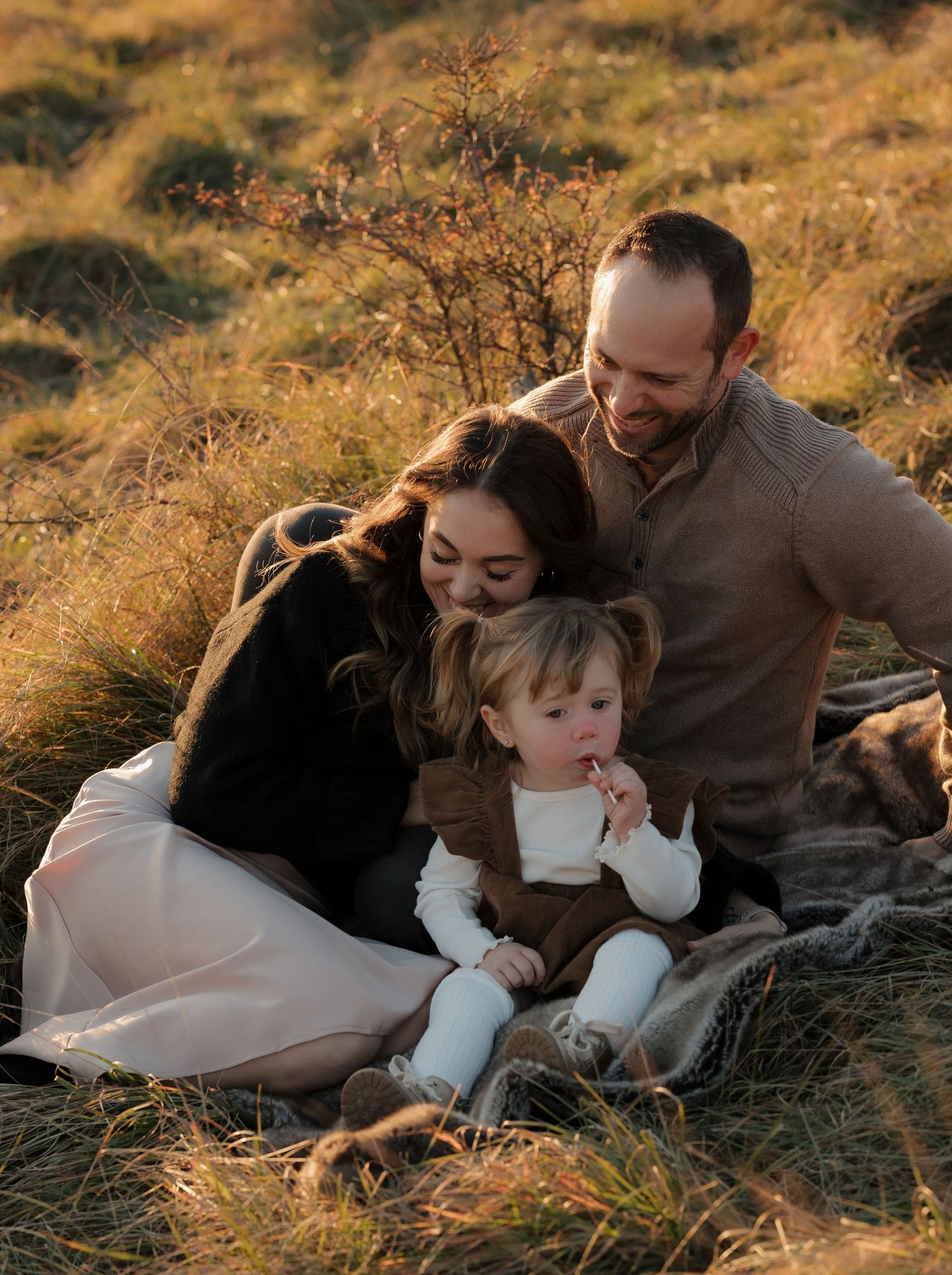 A family lying together on a blanket in tall golden grass during sunset.