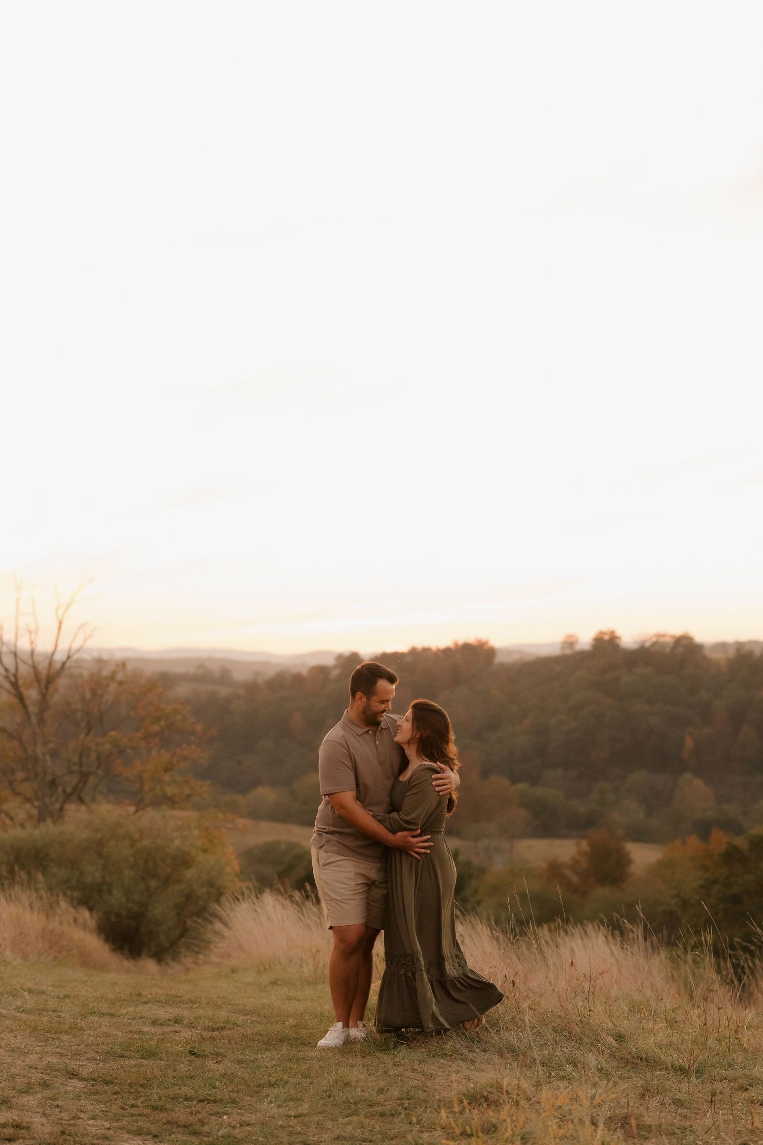 A couple embraces on a hilltop during golden hour sunset with autumn hills in the background.