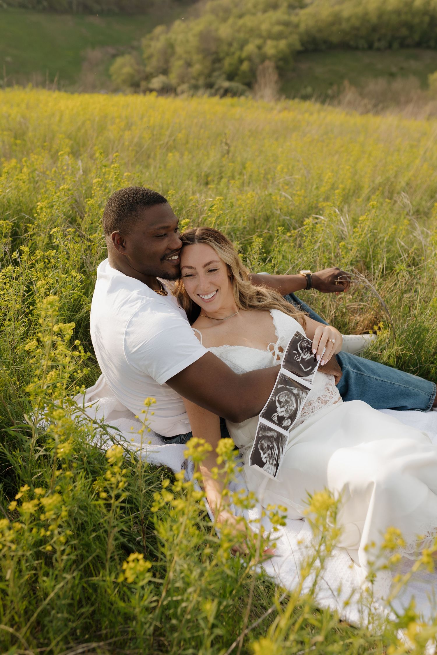 Happy interracial couple sitting in yellow wildflower field, smiling while holding ultrasound photos during outdoor maternity photoshoot.