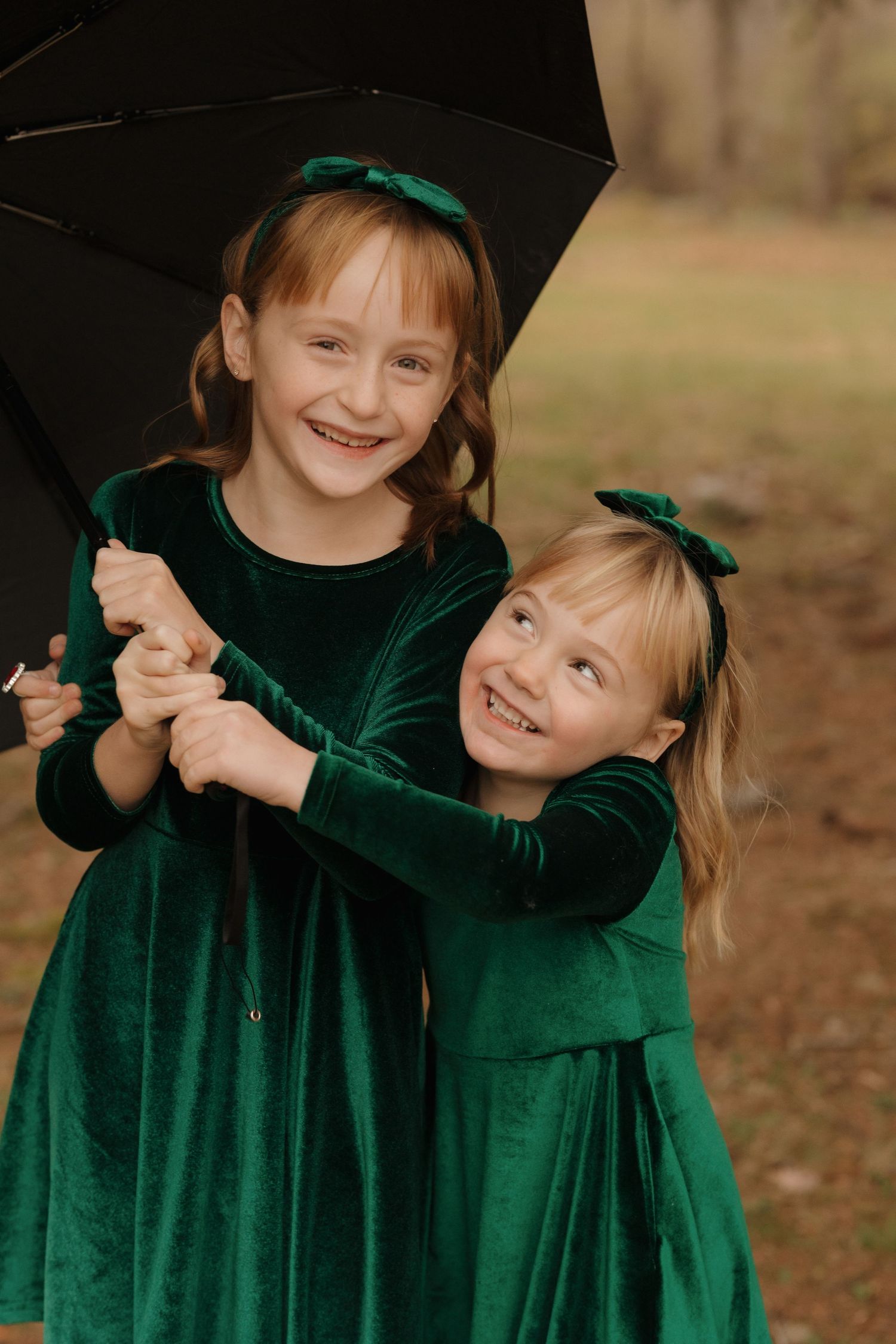 Children in emerald green velvet dresses share joyful moments under a black umbrella.
