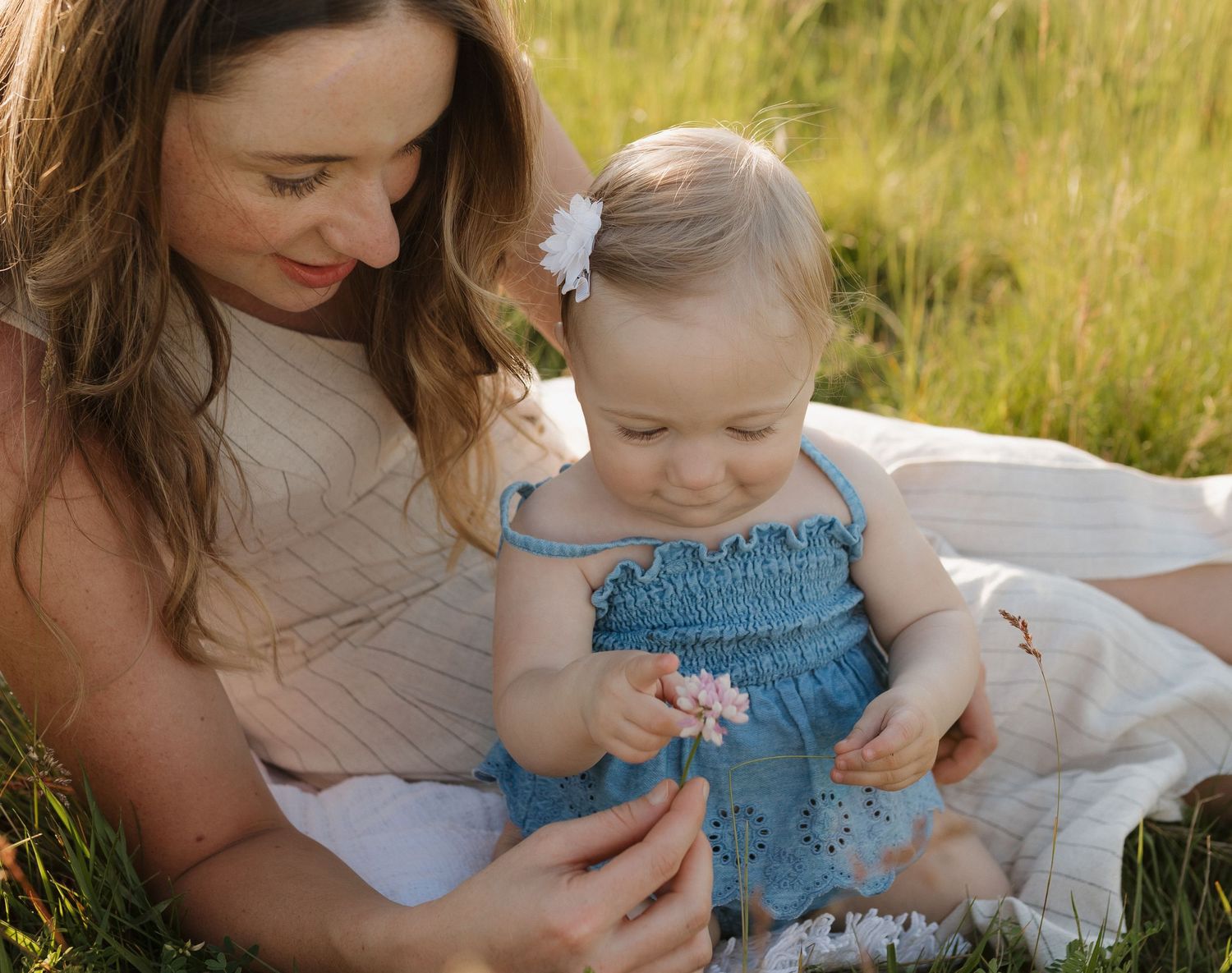 A mother and baby girl in a blue dress sit together in a green meadow, sharing a tender moment outdoors.