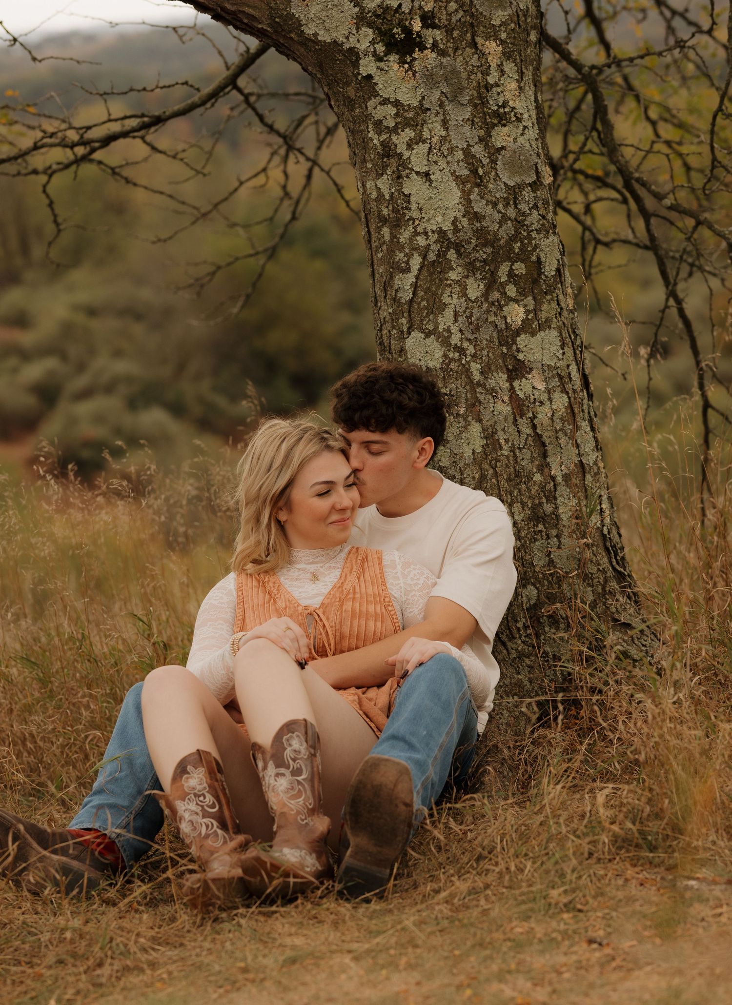 A couple rests beneath a large tree trunk in a rustic outdoor setting.