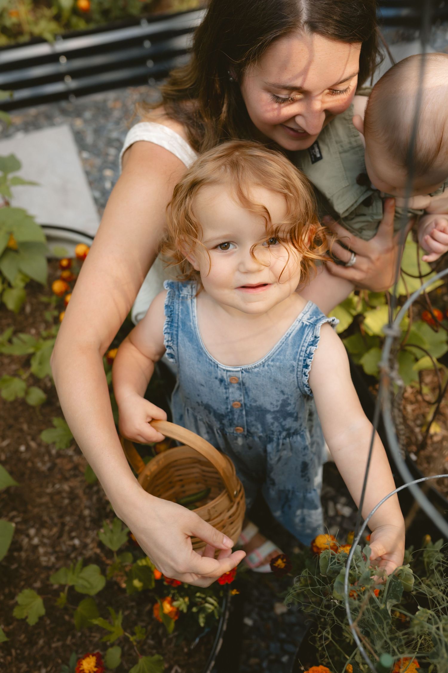 A mother and toddler girl in denim overalls picking vegetables together in a garden, holding a wicker basket.