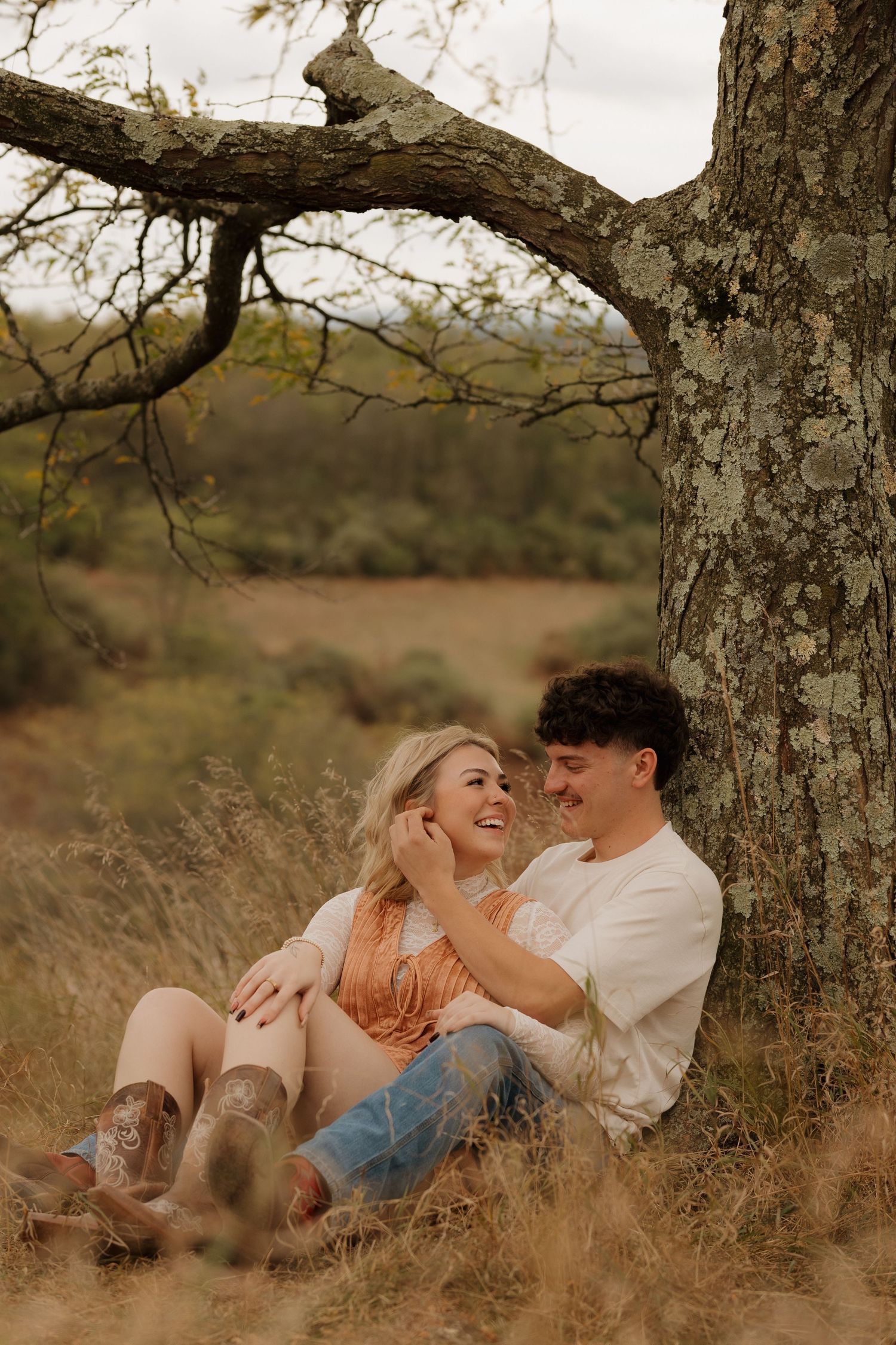 A couple shares intimate moments while lying in tall grass beneath a tree during a romantic outdoor photoshoot.