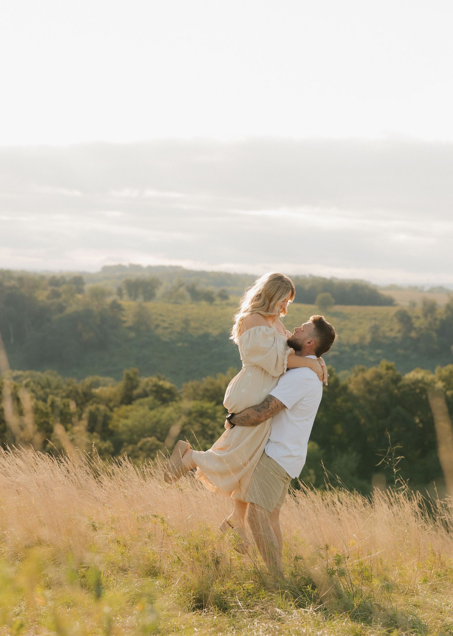 A couple in light colored clothing embrace and twirl together in a sunlit meadow of tall grass against a rolling landscape.