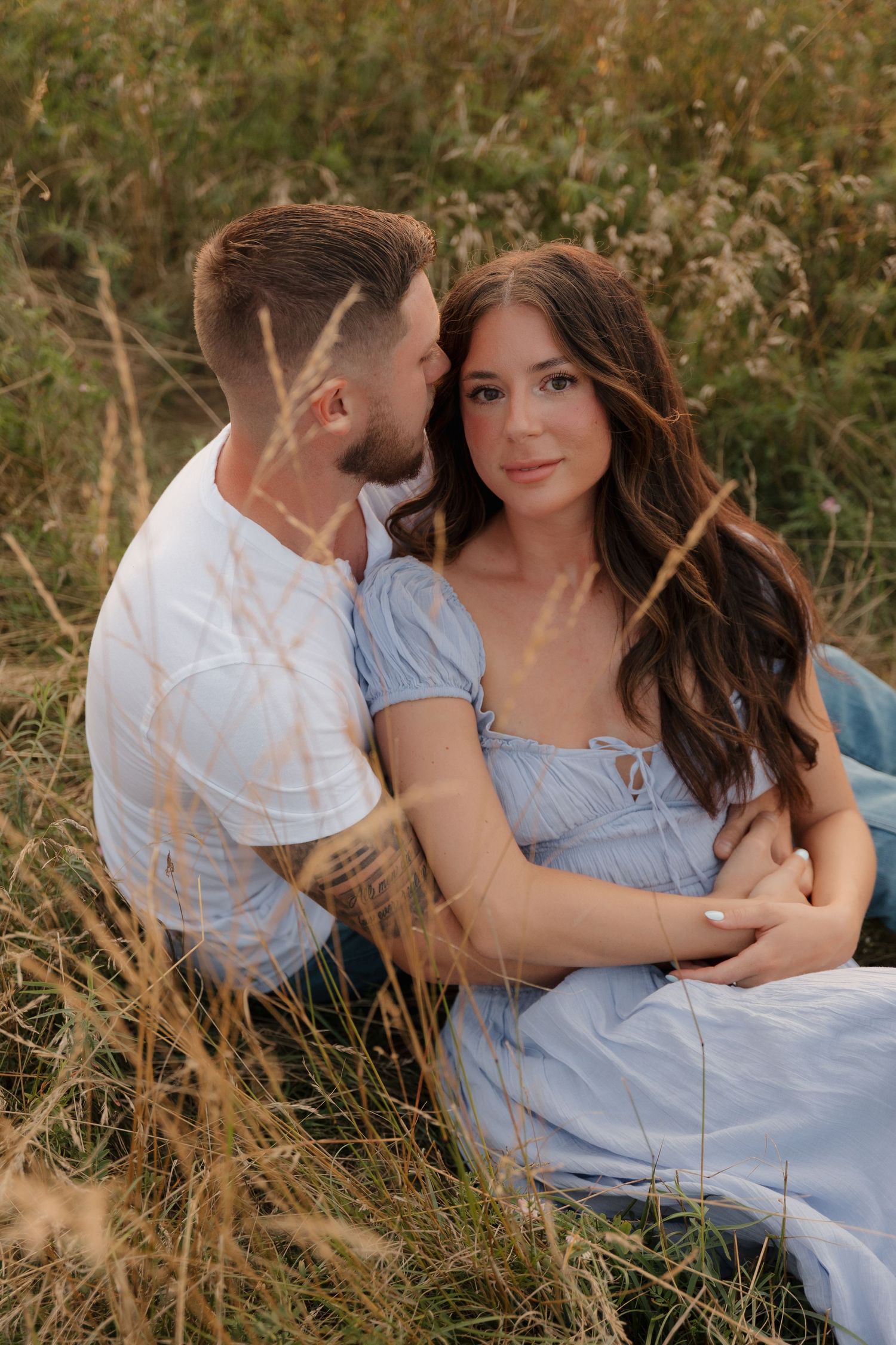 Couple sharing tender moments while sitting in tall grass during a countryside photoshoot.