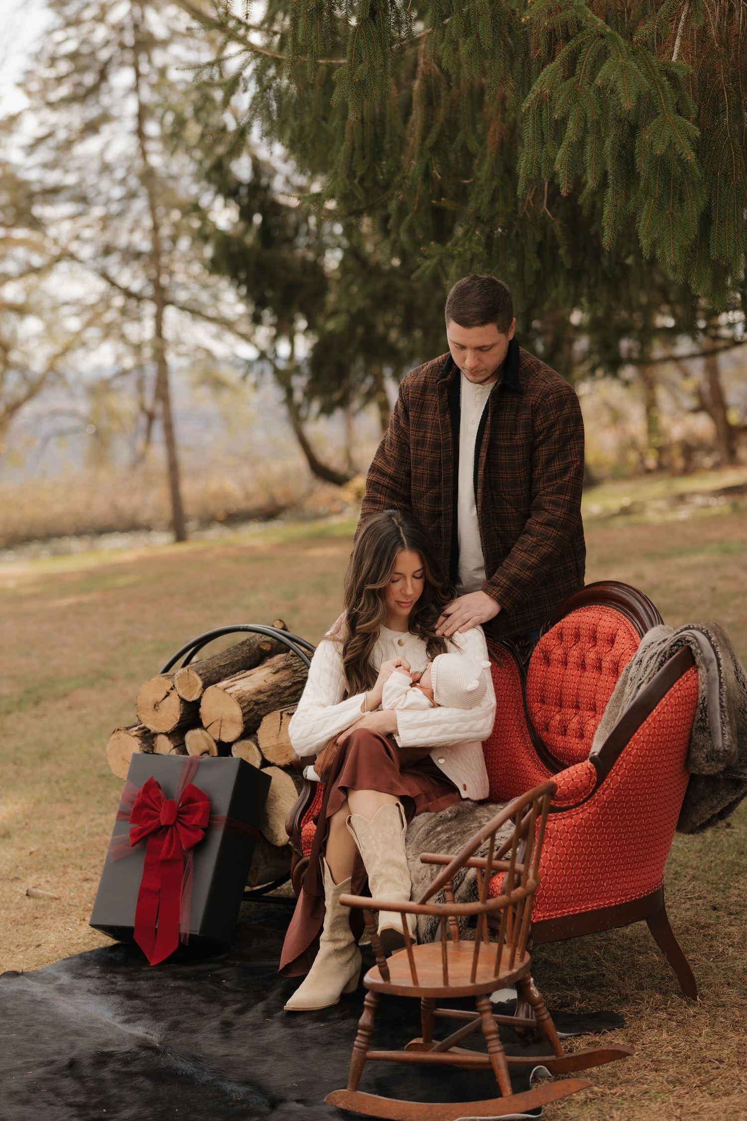Family relaxes together on a red vintage settee outdoors under pine trees.