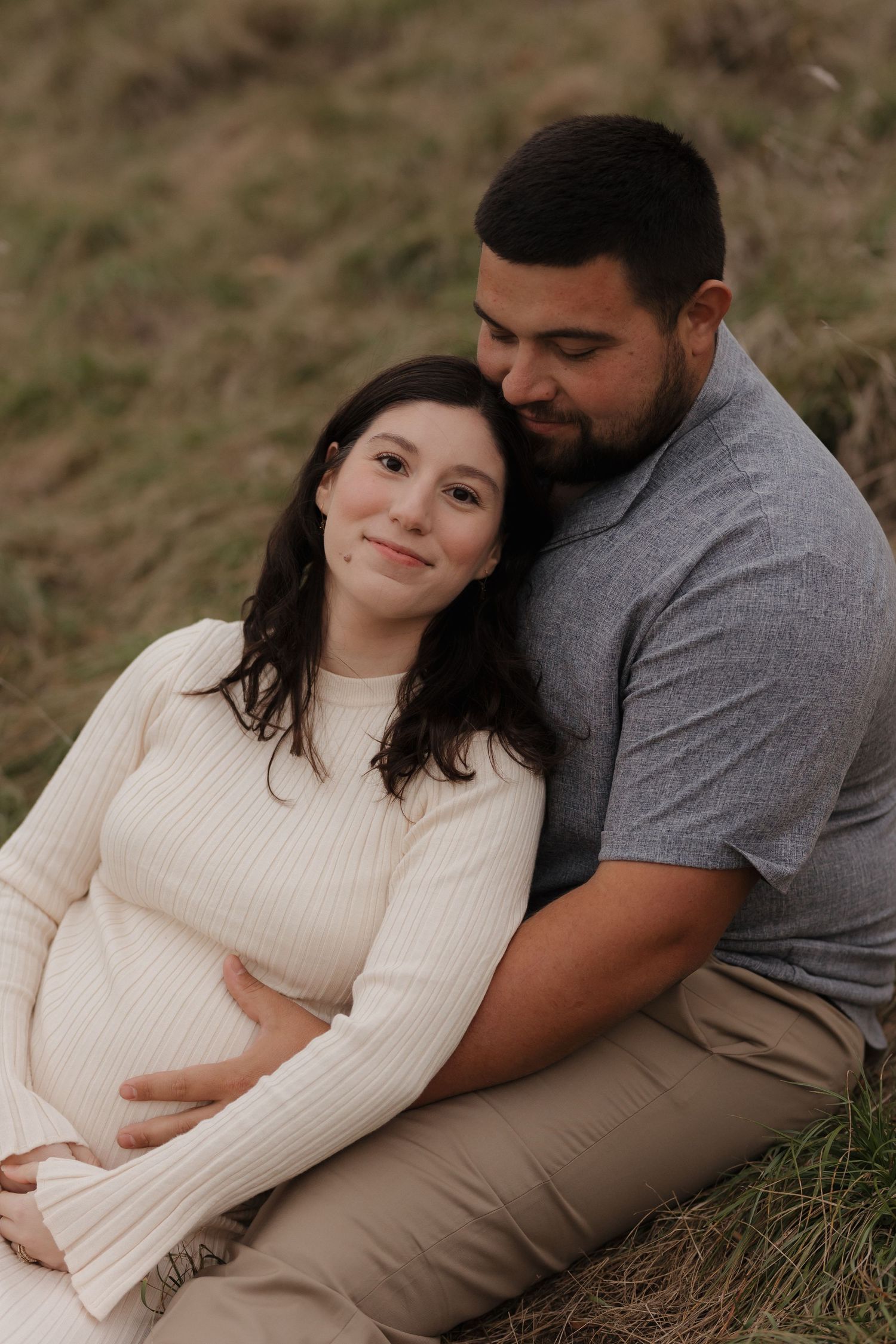 A couple cuddles together on grassy ground during an outdoor photo session.