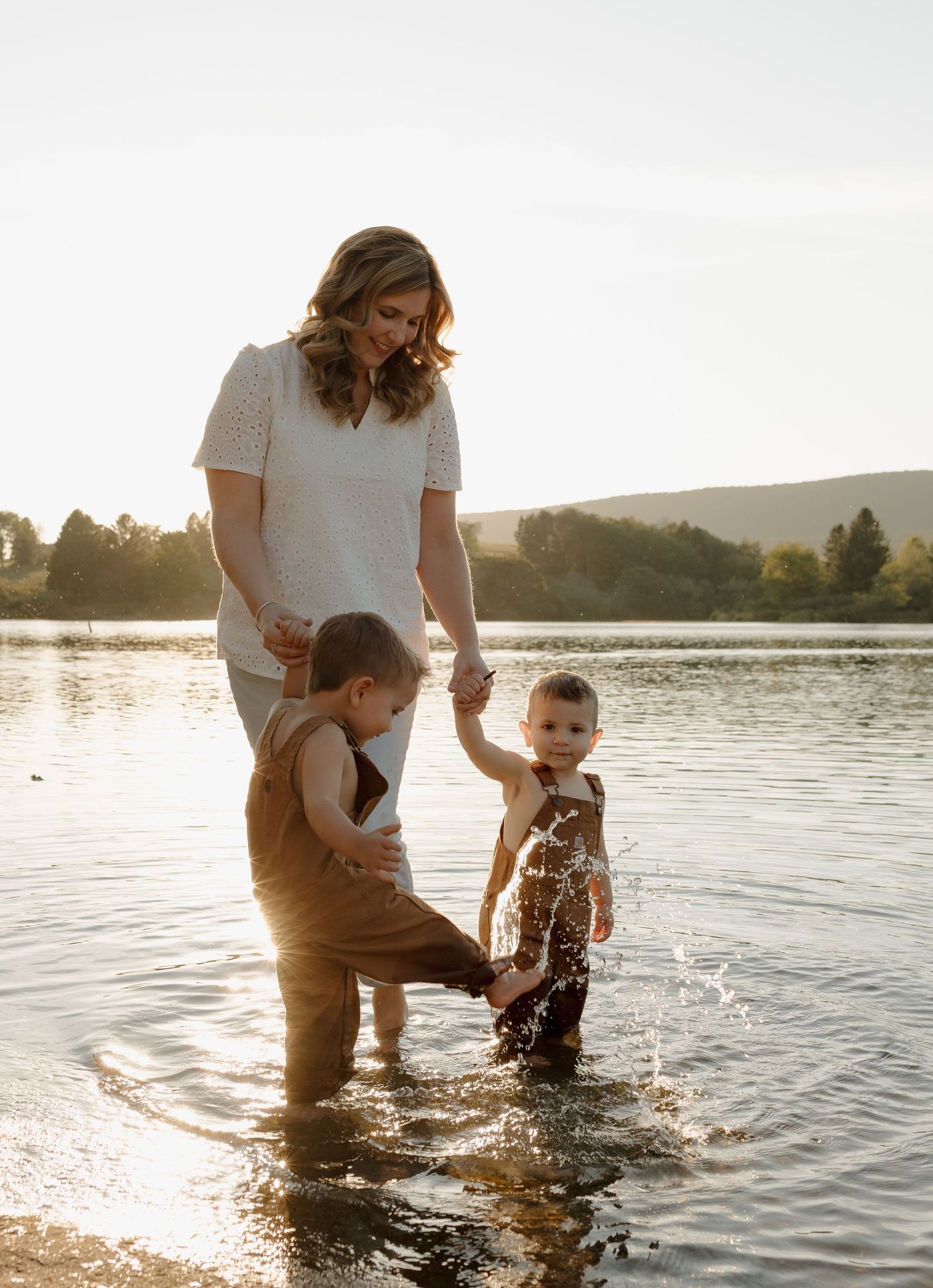 A mother and two toddlers play in a sunlit lake, holding hands and splashing water during golden hour.