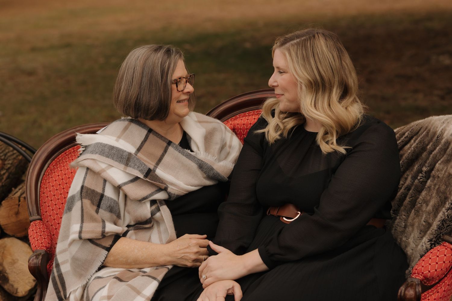 Two women share a tender moment while sitting on a vintage red settee outdoors.