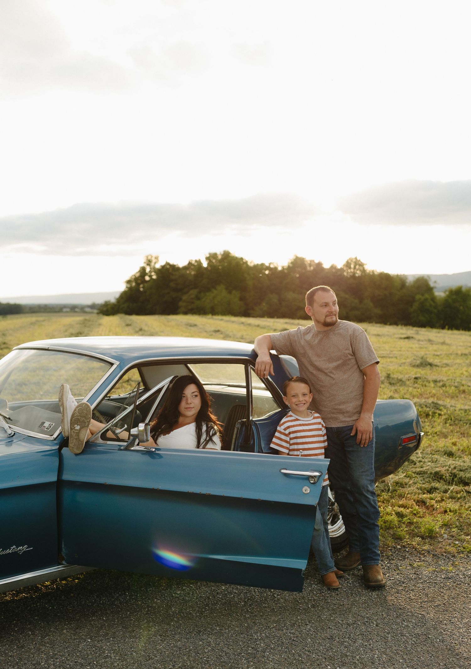 Family posing with a vintage teal classic car in a scenic rural field at golden hour sunset.