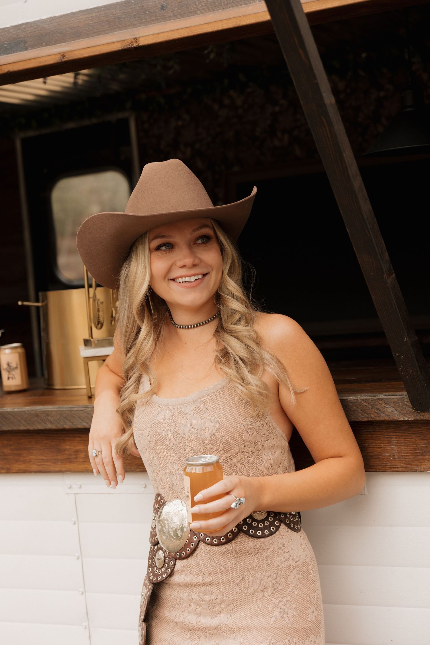 A woman in a beige dress and cowboy hat holds a drink at a rustic bar.