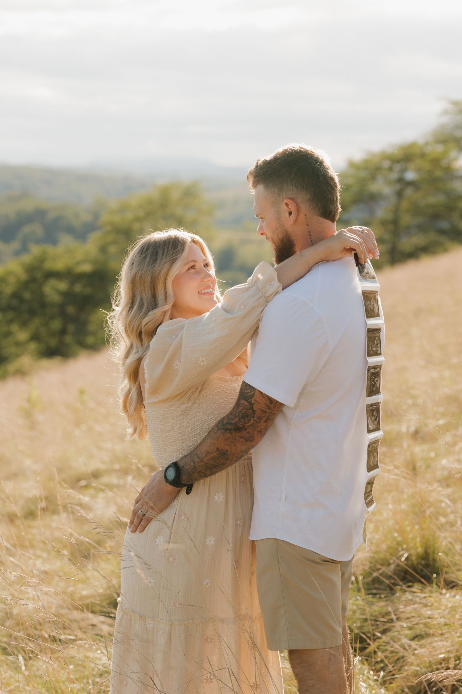 A couple embraces in a golden field at sunset, with rolling hills and trees visible in the background.