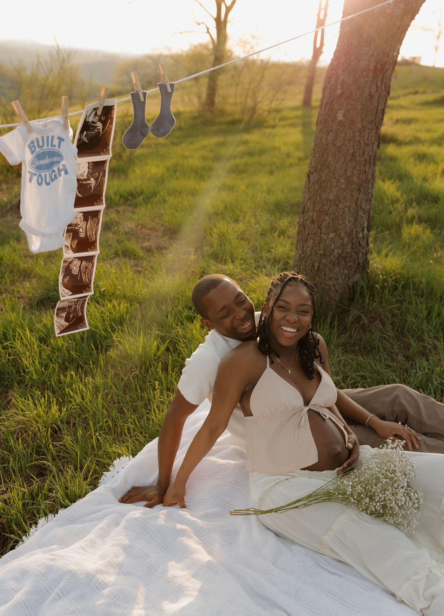 Couple sitting on white blanket outdoors at golden hour with baby ultrasound photos and onesie on clothesline.
