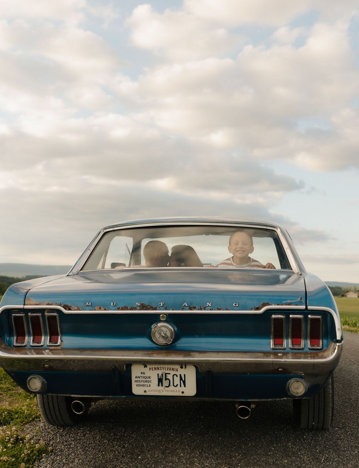 Rear view of a classic blue Ford Mustang with Pennsylvania plates on a rural road, passenger visible through rear window.