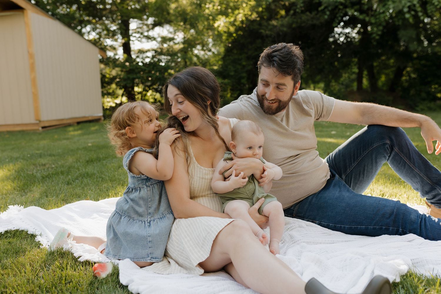 Happy family of four sitting on a white blanket outdoors, parents laughing with toddler and baby in a lush green backyard.